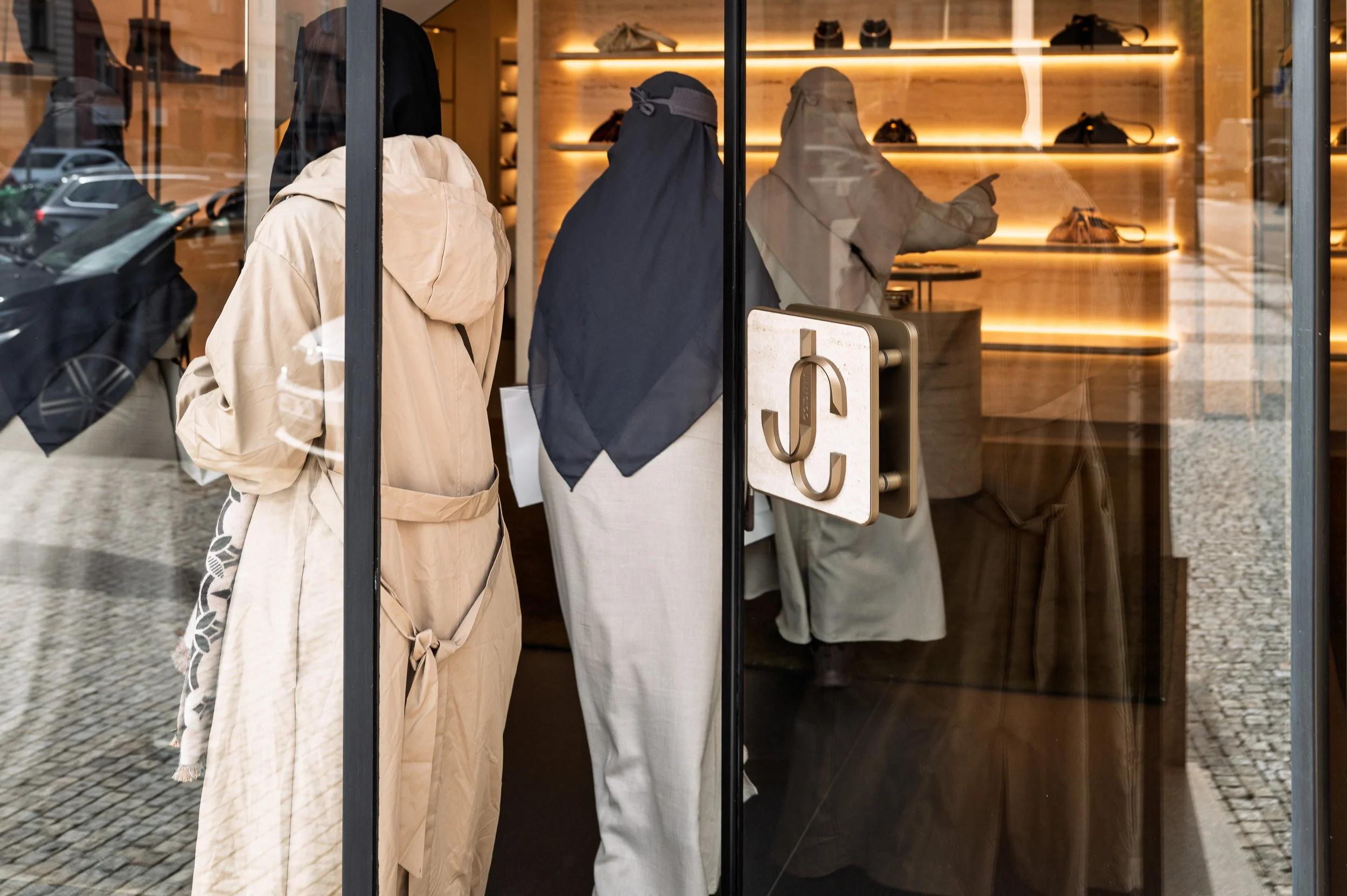 Two women in long robes at a boutique door, a third inside among handbags, street reflected, Prague — Leica Q3