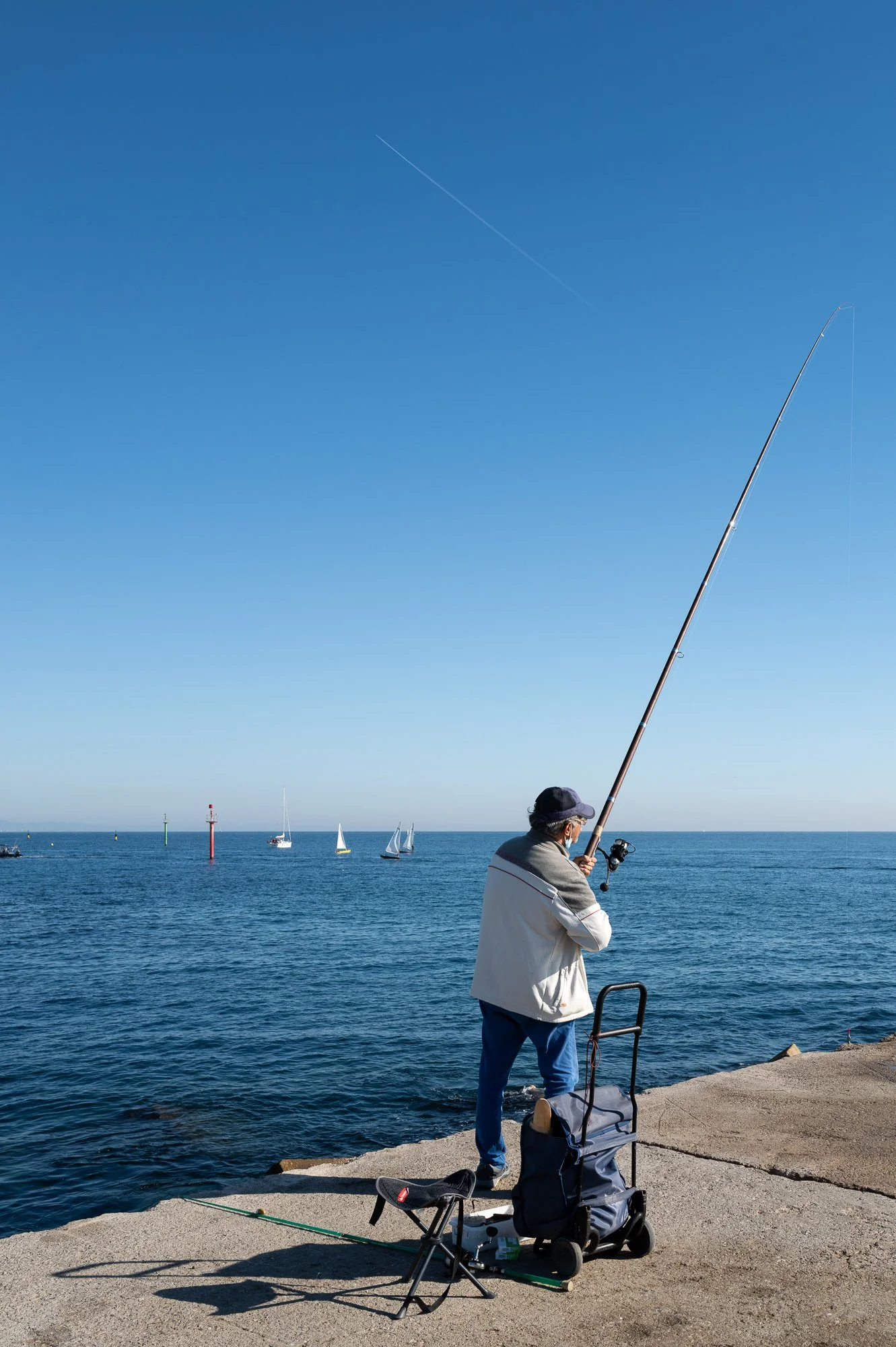 Fisherman with a long rod on the breakwater, contrail and sailboats behind, Barceloneta — Leica Q3