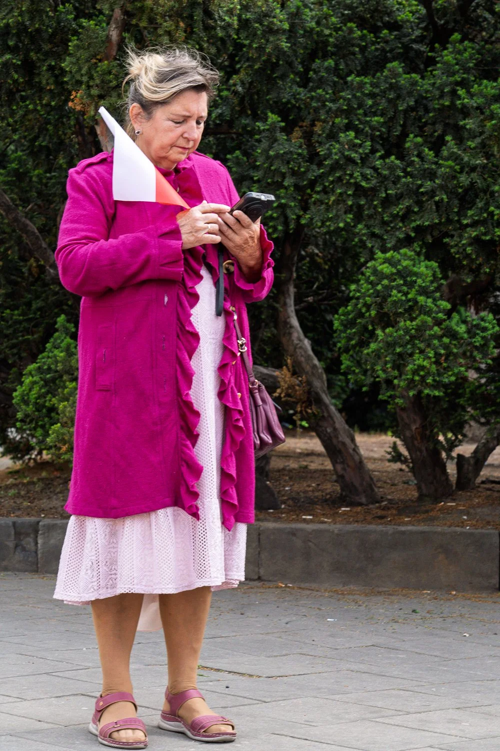 Older woman in a magenta ruffled coat holding a small Polish flag checks her phone, Warsaw — Sony a7R