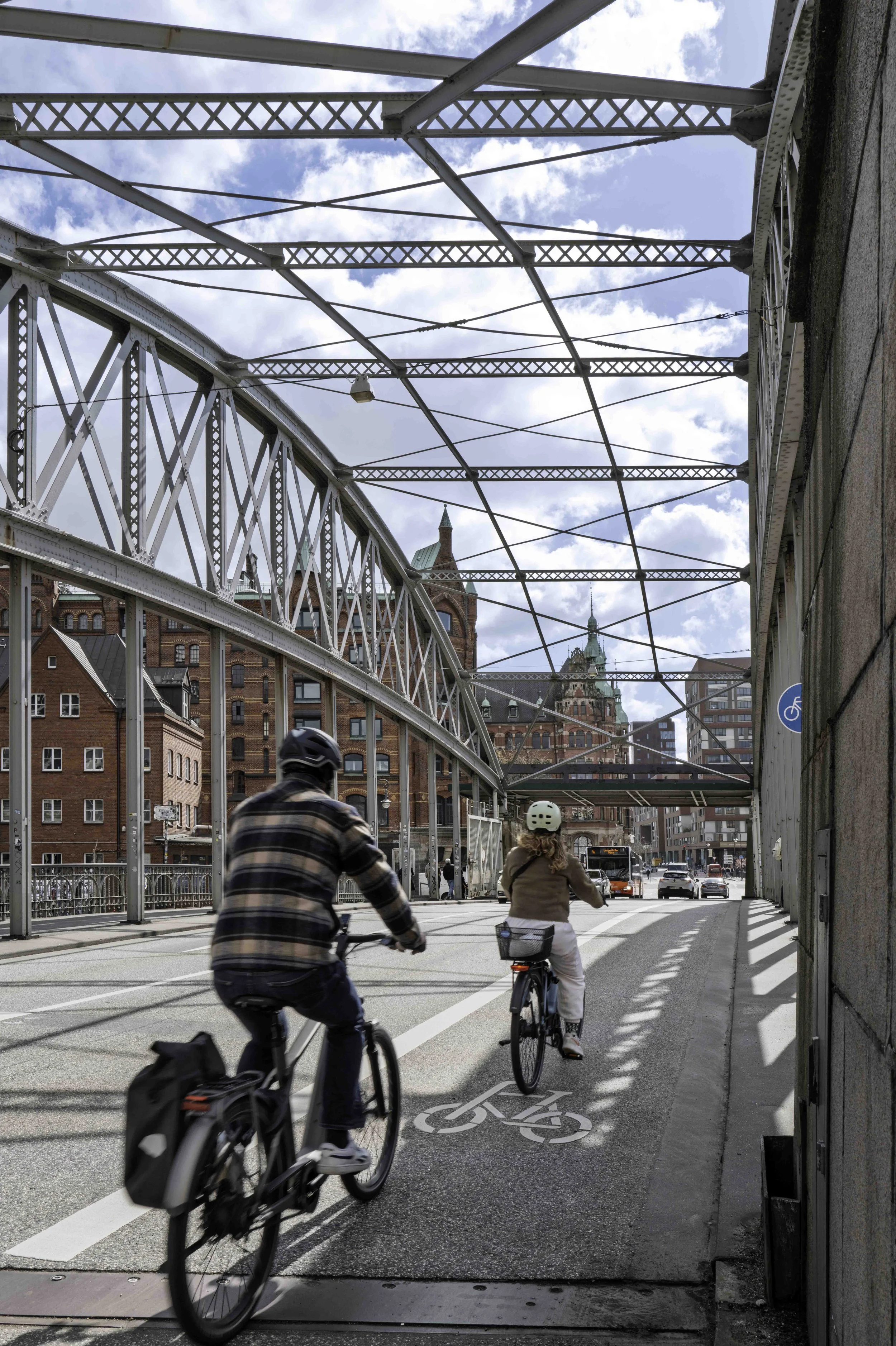 Two cyclists riding through steel truss bridge, Speicherstadt red-brick warehouses visible through the arch, Hamburg — Leica Q3