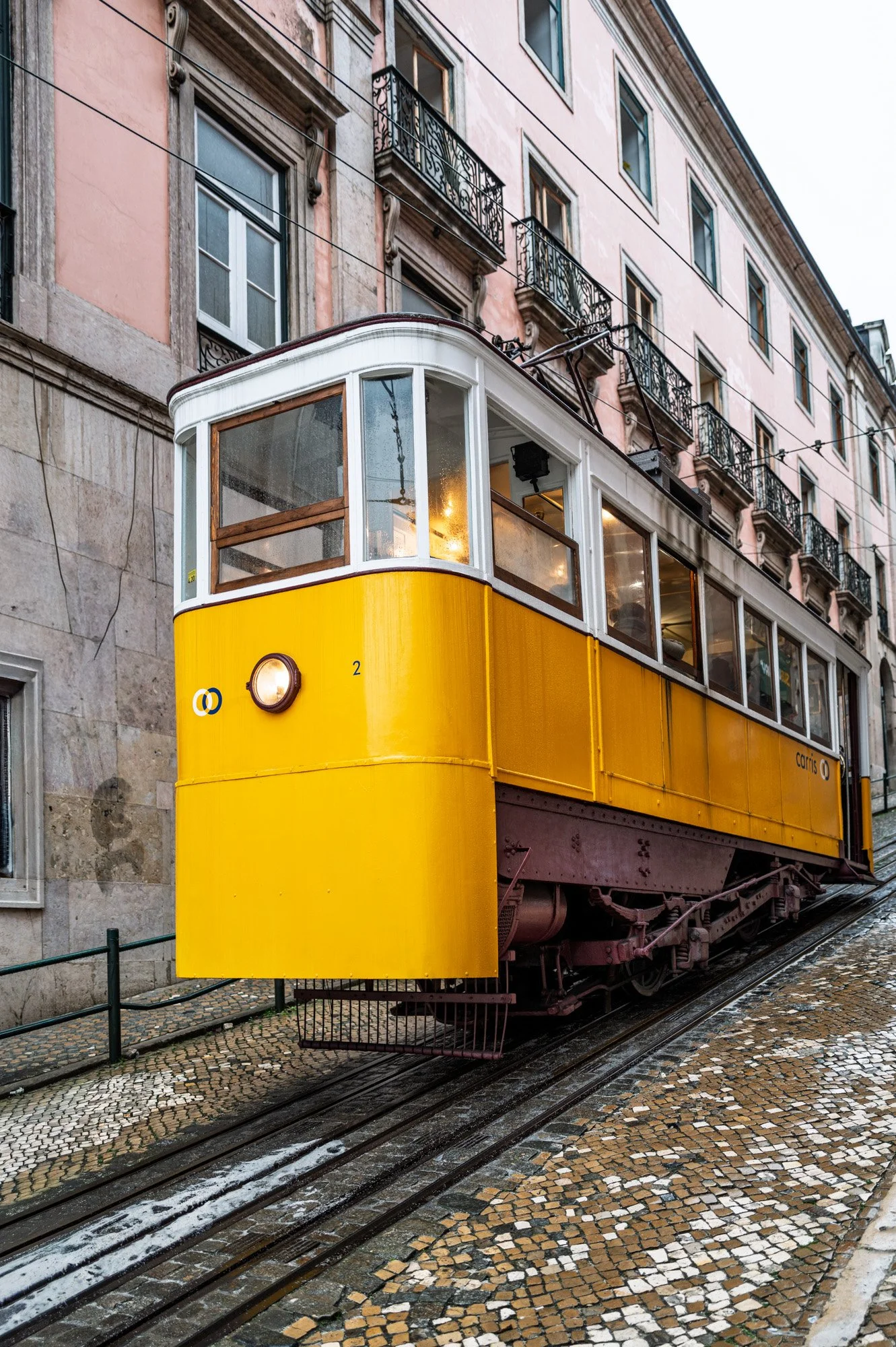 Yellow Glória funicular tram on a wet cobblestone street with pink building facade, Lisbon — bCLPhoto