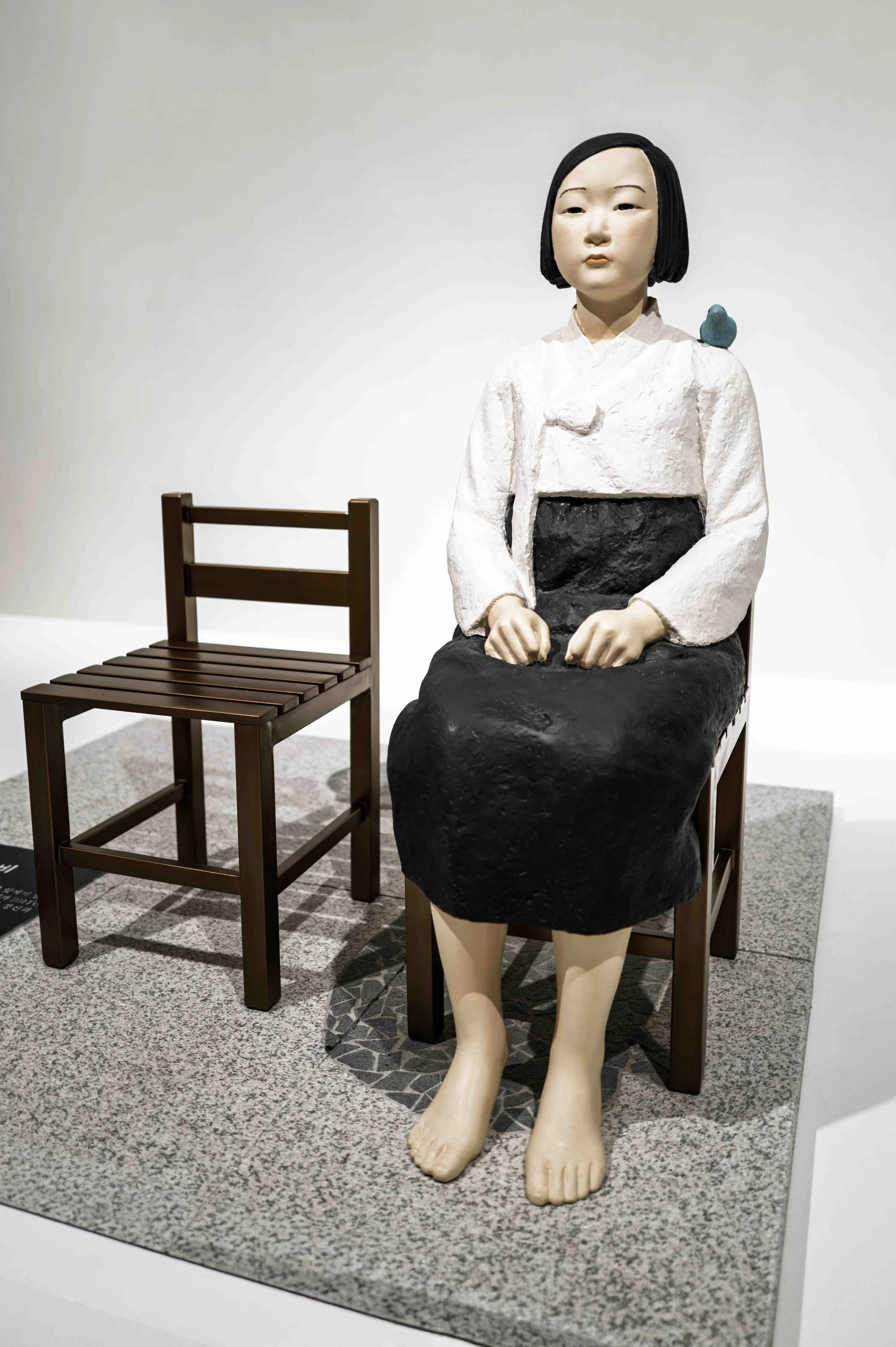 Painted statue of a barefoot girl in a white jacket and black skirt seated on a wooden chair, an empty chair beside her