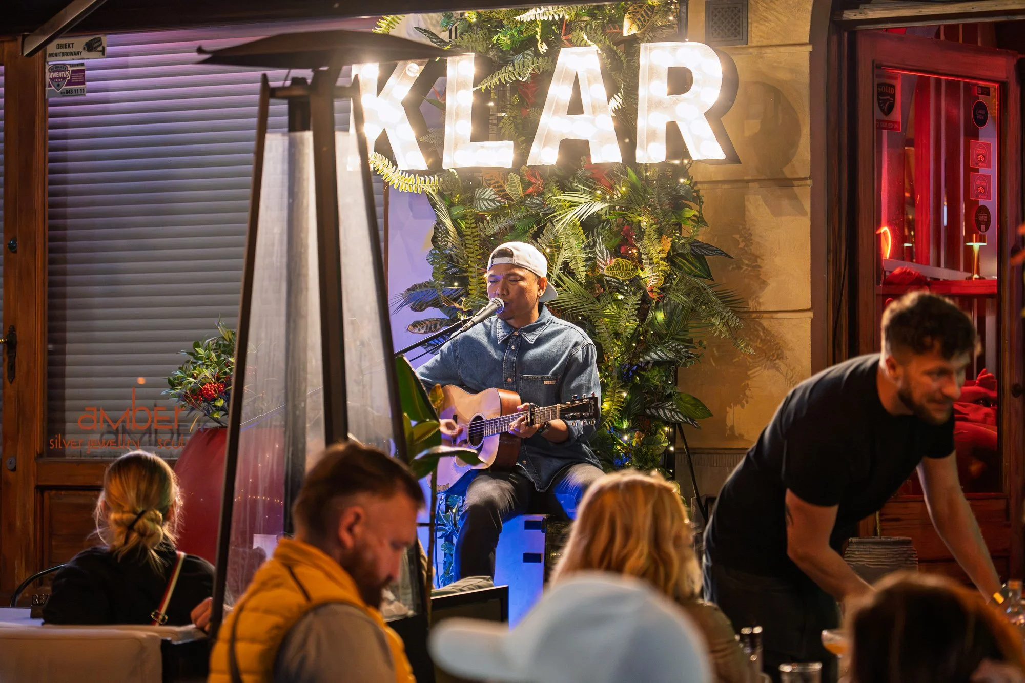 Street musician with a guitar under illuminated KLAR letters while diners talk and a waiter passes, Warsaw — Sony a7R IV