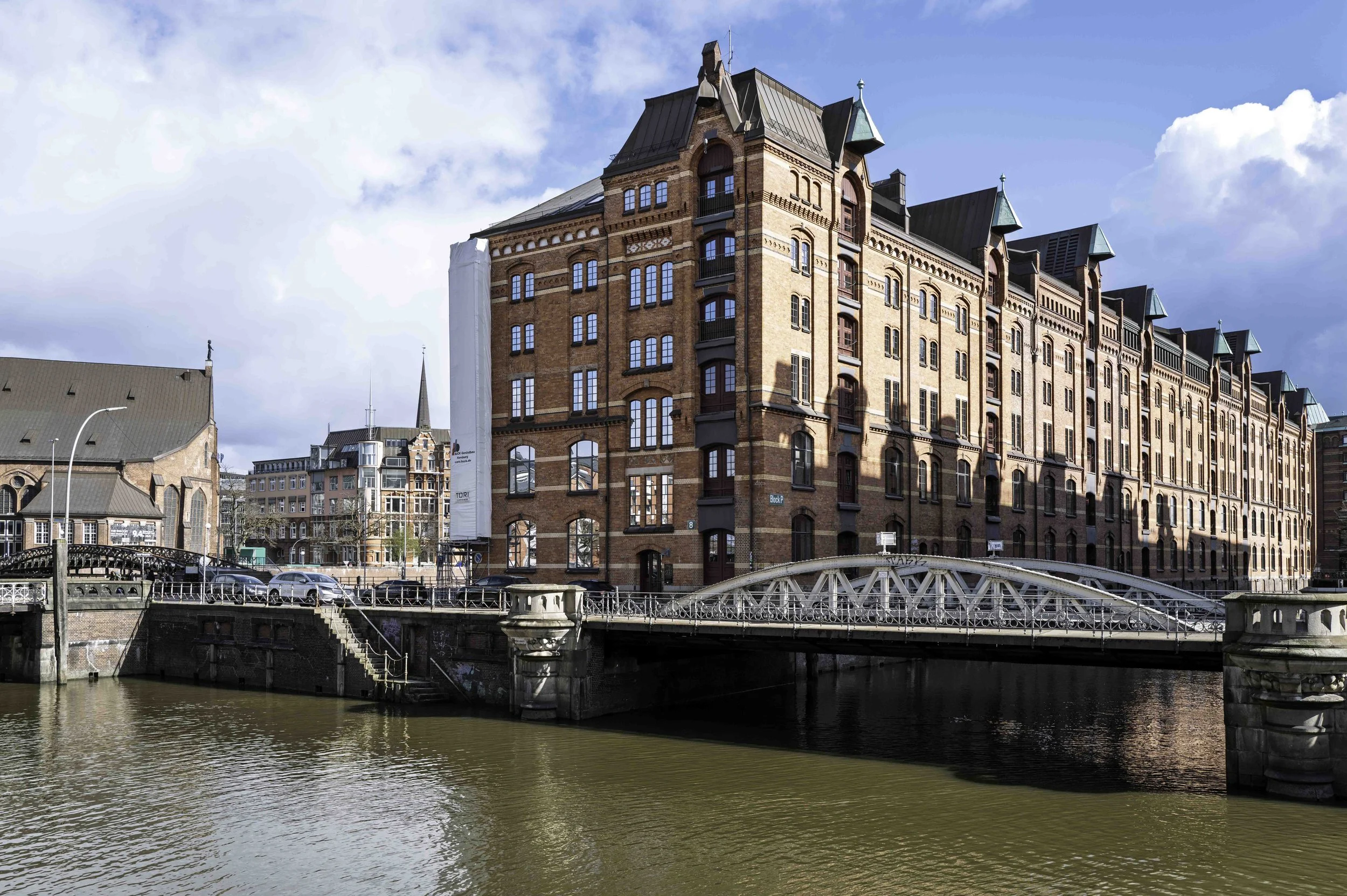 Low iron bridge in foreground, sunlit red-brick Speicherstadt warehouses stretching behind, Hamburg — Leica Q3