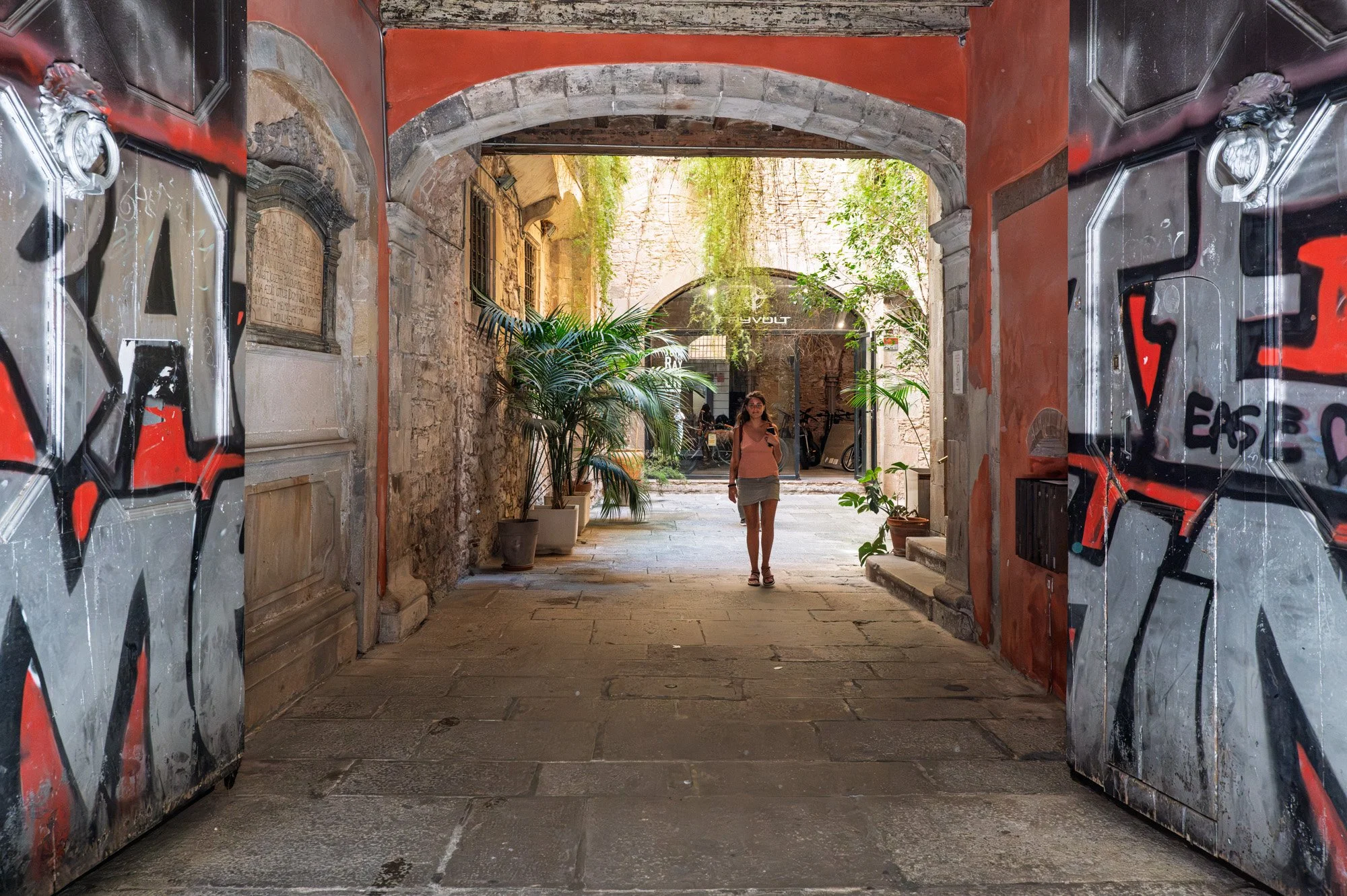 Woman walking through graffiti-covered doors into a sunlit courtyard, El Born — Leica Q3