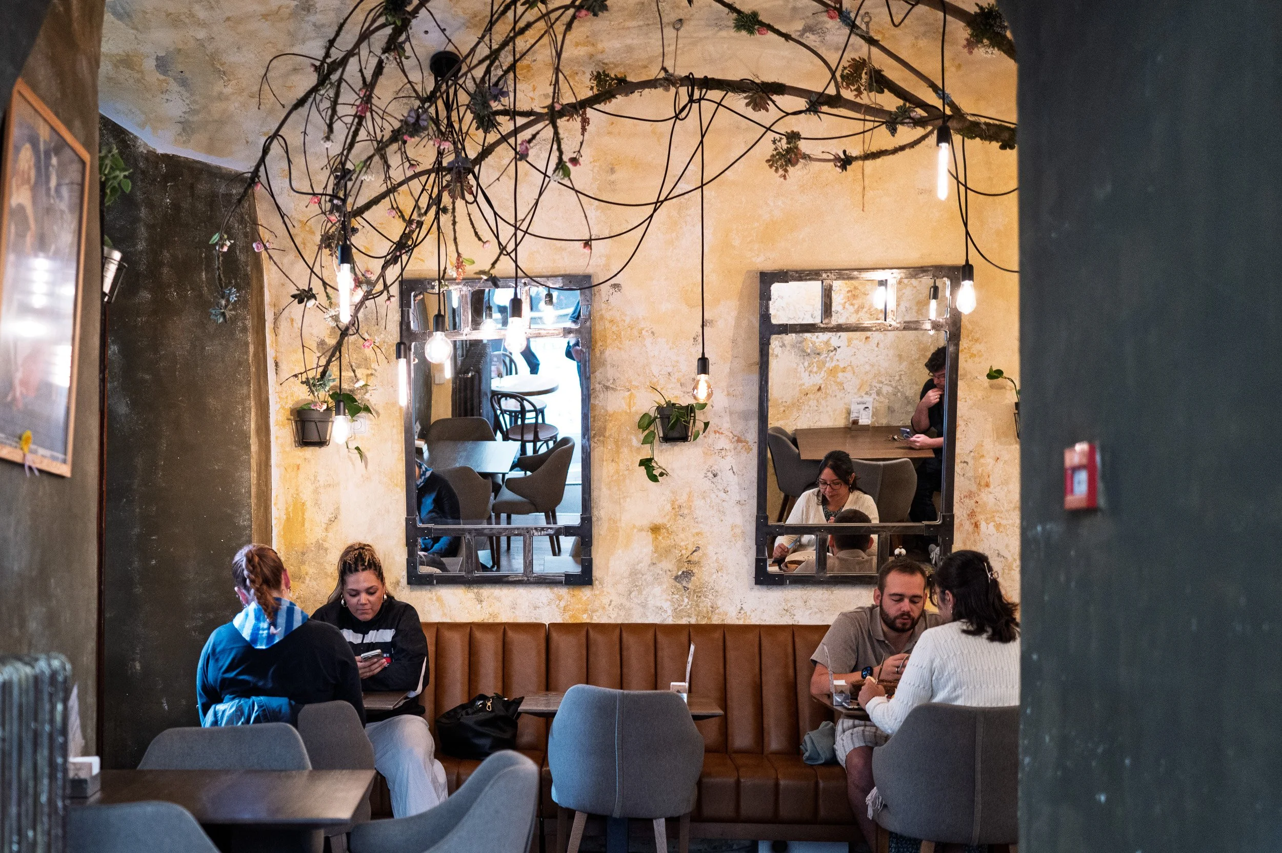 Café interior with two iron-framed mirrors, hanging branches and bulbs, diners below, Prague — Leica Q3