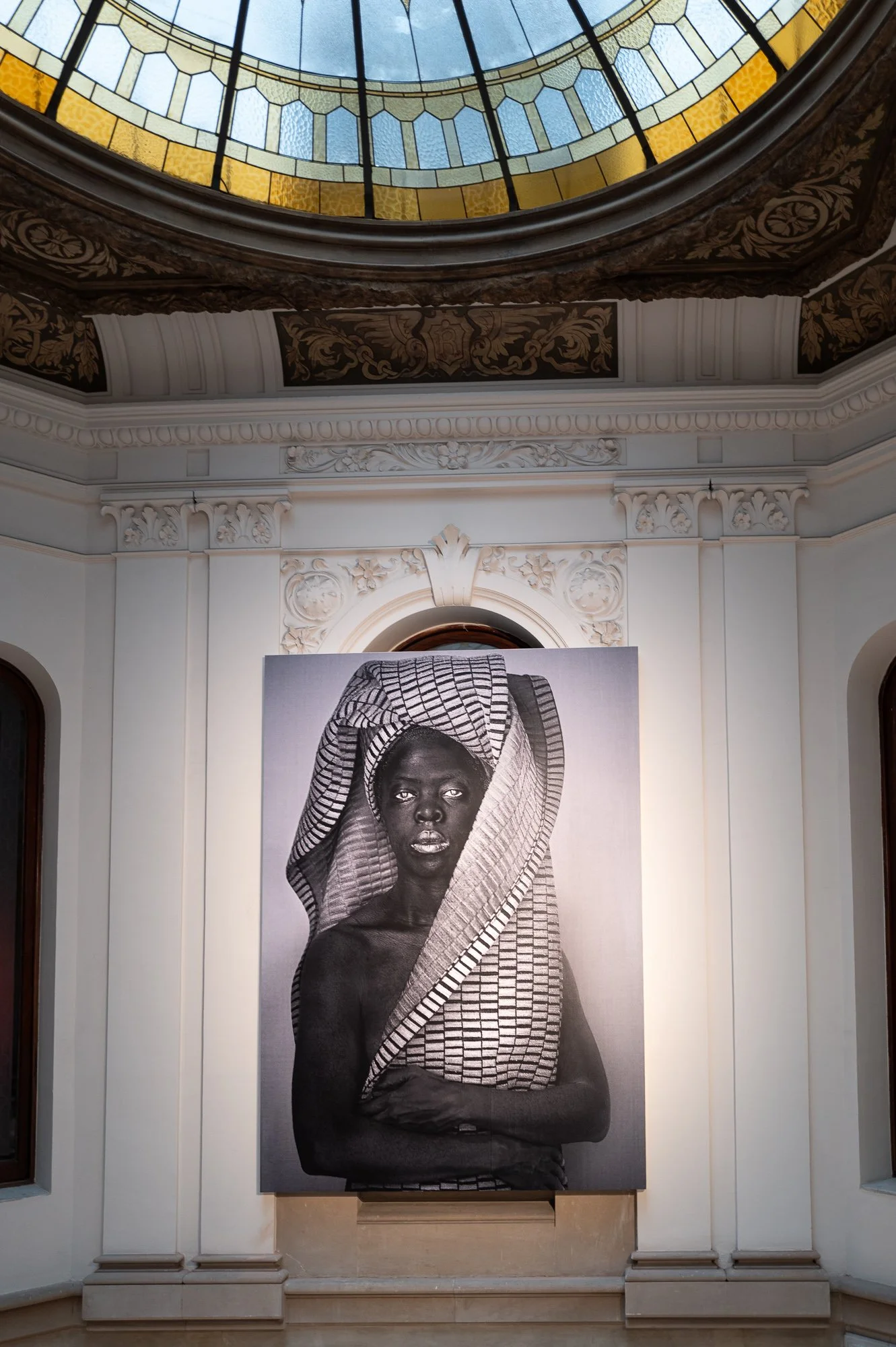 A large black and white portrait hangs below an ornate stained-glass skylight with gilded mouldings in a white stairwell