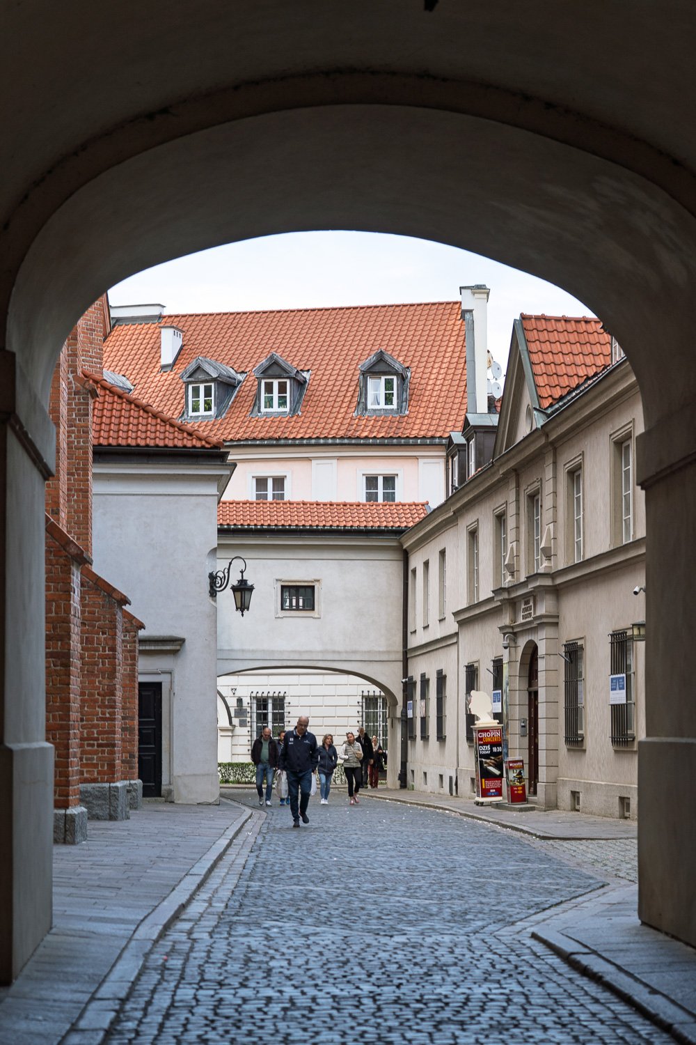 Stone archway framing red-tiled roofs and a cobbled street in Warsaw Old Town, people walking in — Sony a7R IV