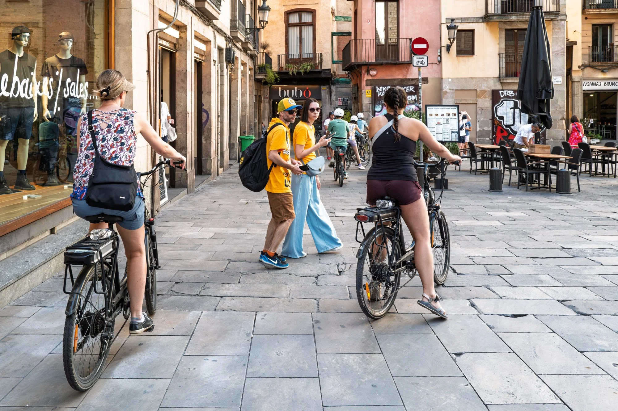 Cyclists in yellow shirts weaving past pedestrians near Bubó, El Born — Leica Q3