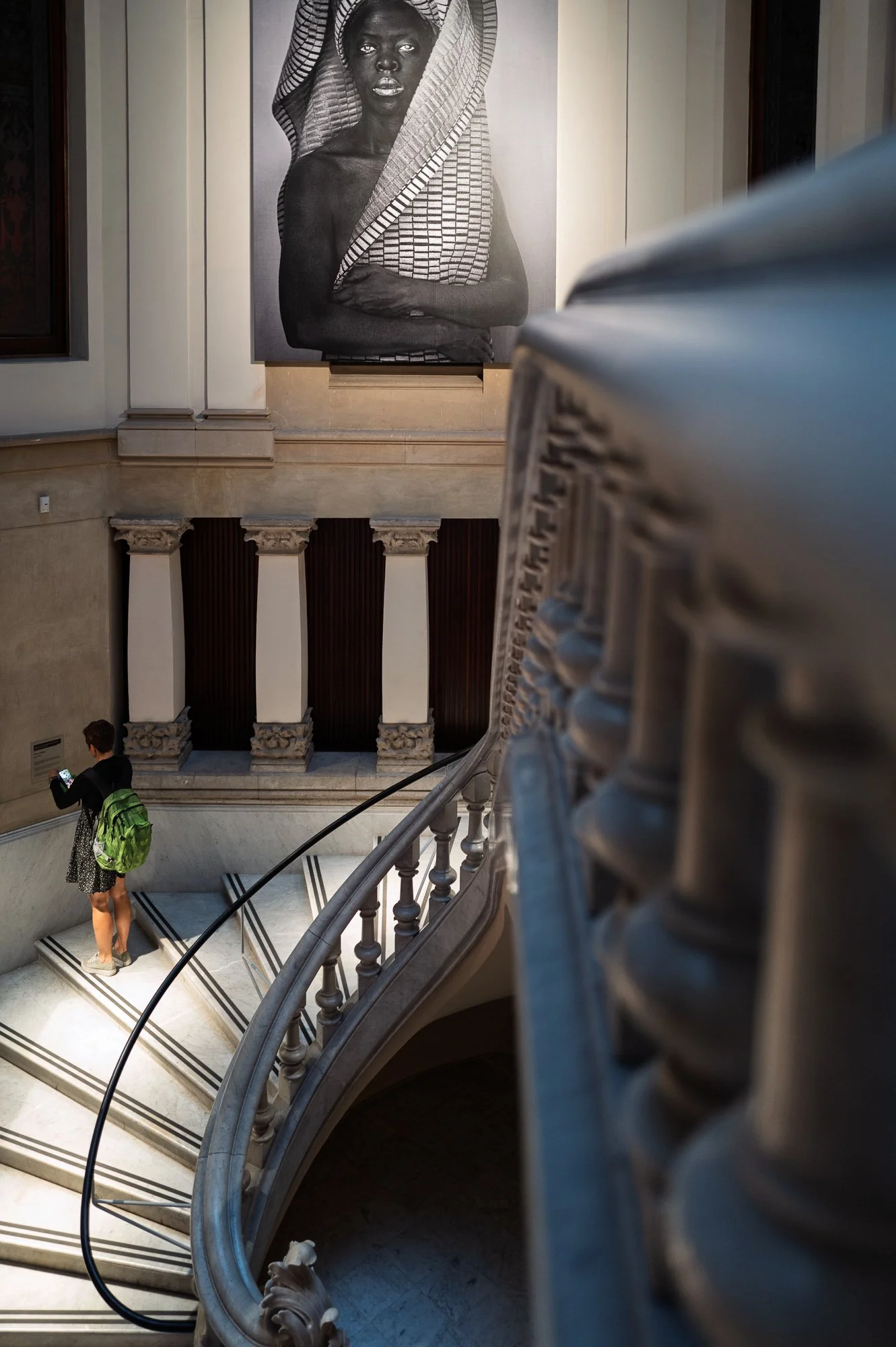 A young woman with a green backpack photographs a wall label with her phone on the staircase, the balustrade blurred in front