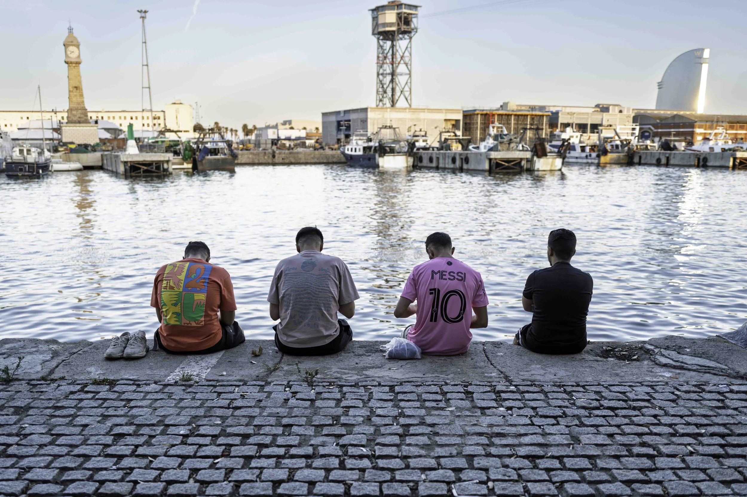 Four young men sitting with their backs to the camera looking out over Barcelona port at dusk, one wearing a Messi 10 Inter Miami jersey — Leica Q3
