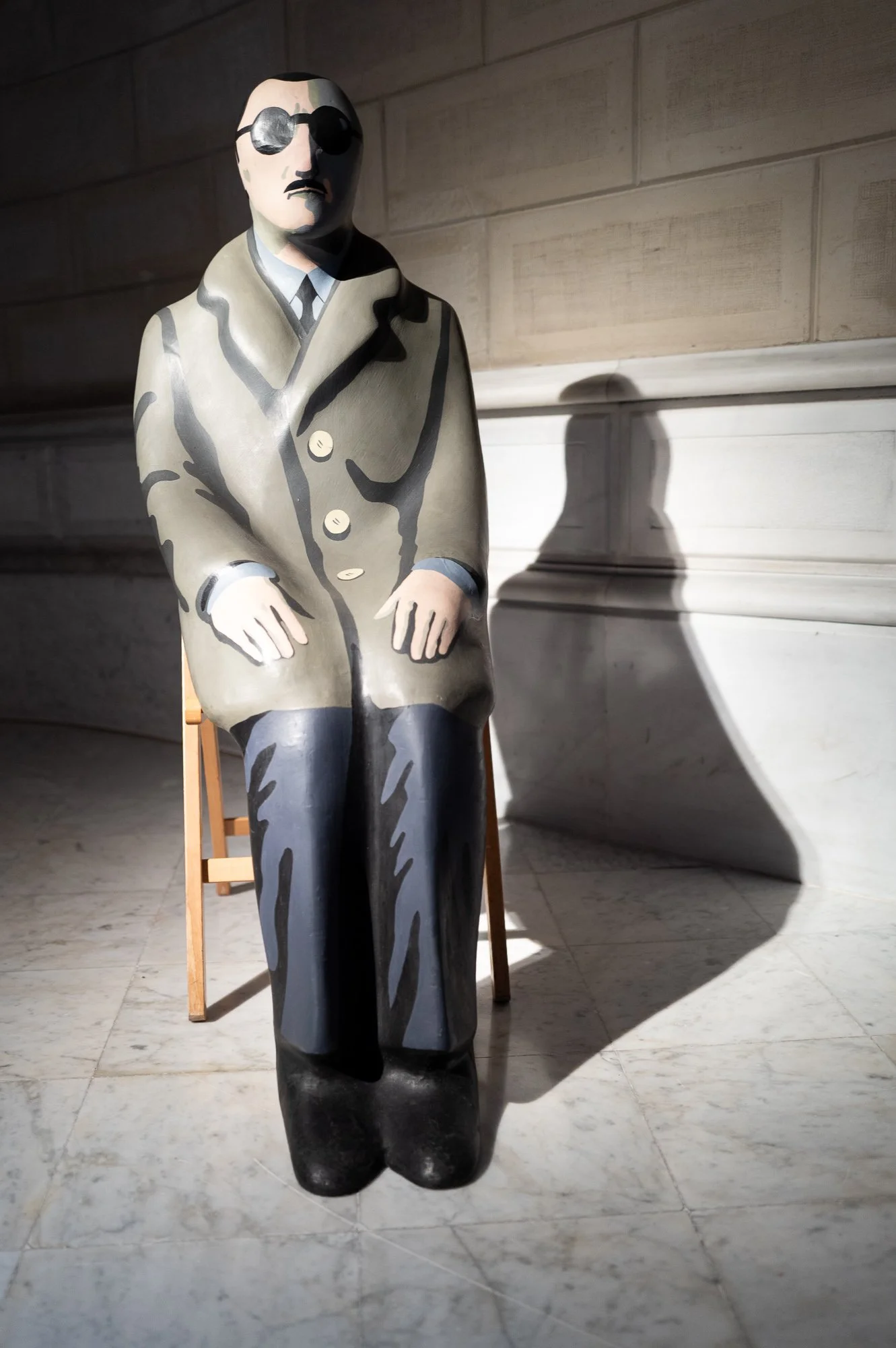 Painted sculpture of a man in a grey overcoat, black glasses and moustache, seated on a folding chair, his shadow on the wall