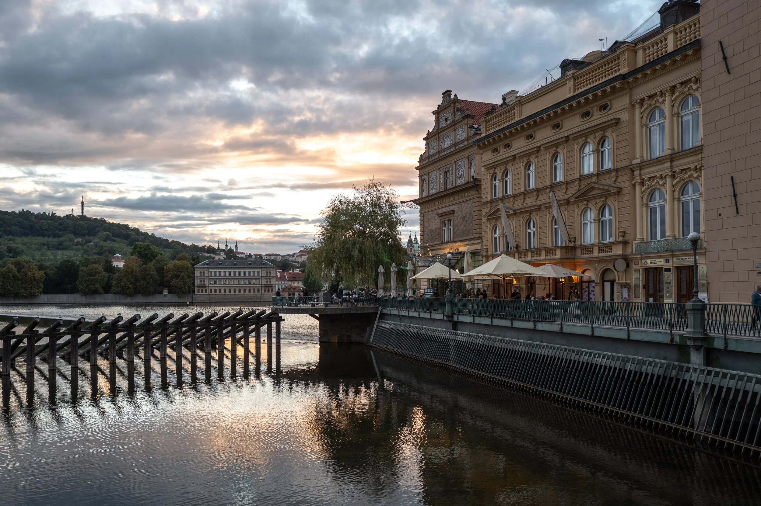 Sunset over Petřín Hill, timber posts reflected in the Vltava by the Lávka terraces, Prague — Leica Q3