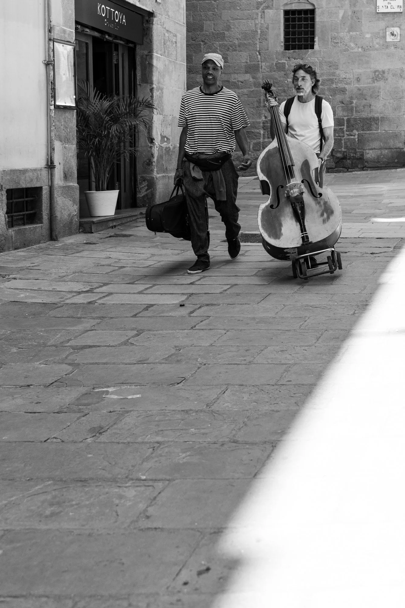 Two musicians walking through a Gothic Quarter street, one pulling a double bass on a trolley, Barcelona — Leica Q3