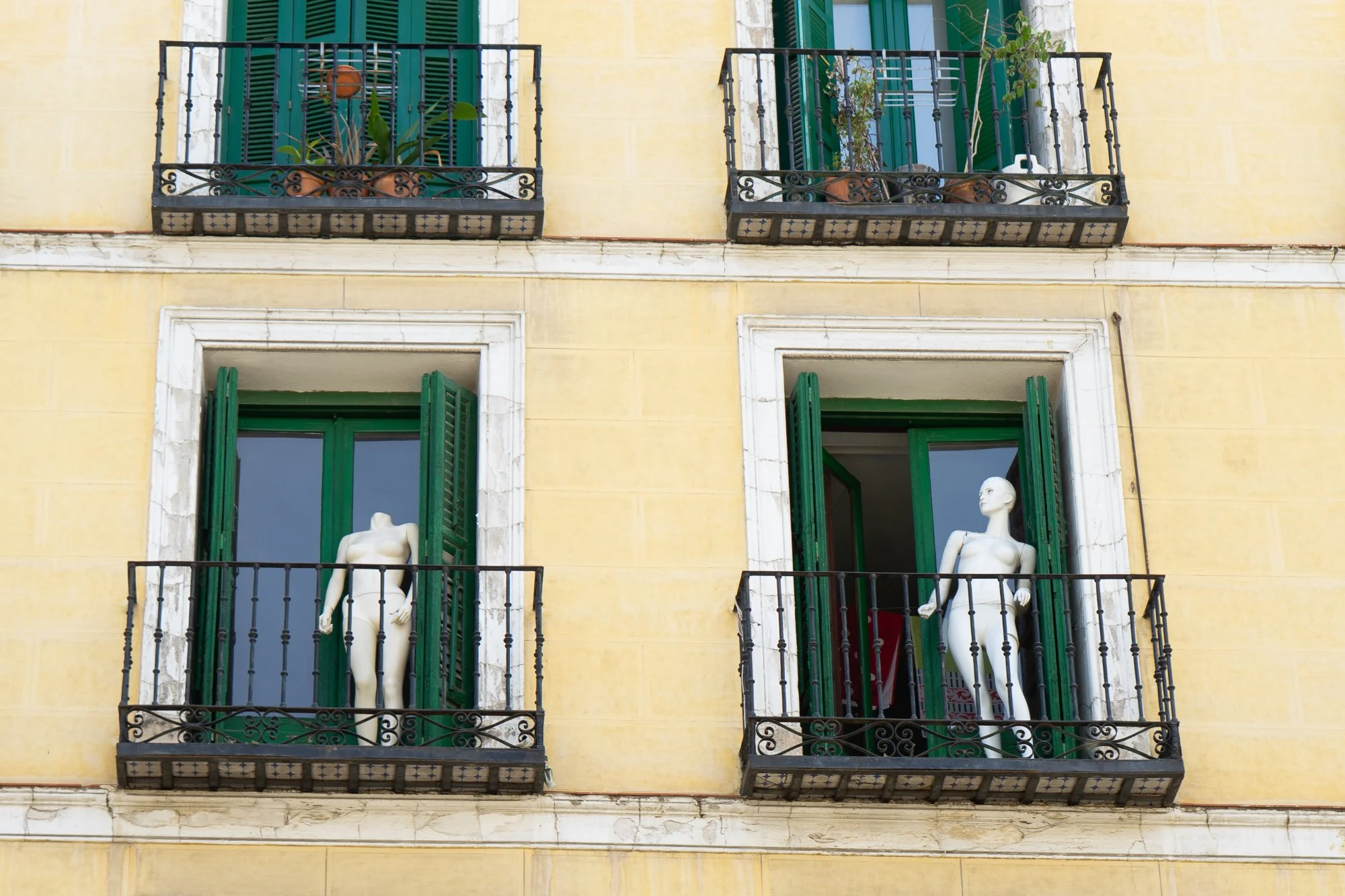 Two white mannequins stand in open balcony windows with green shutters on a yellow façade, Madrid