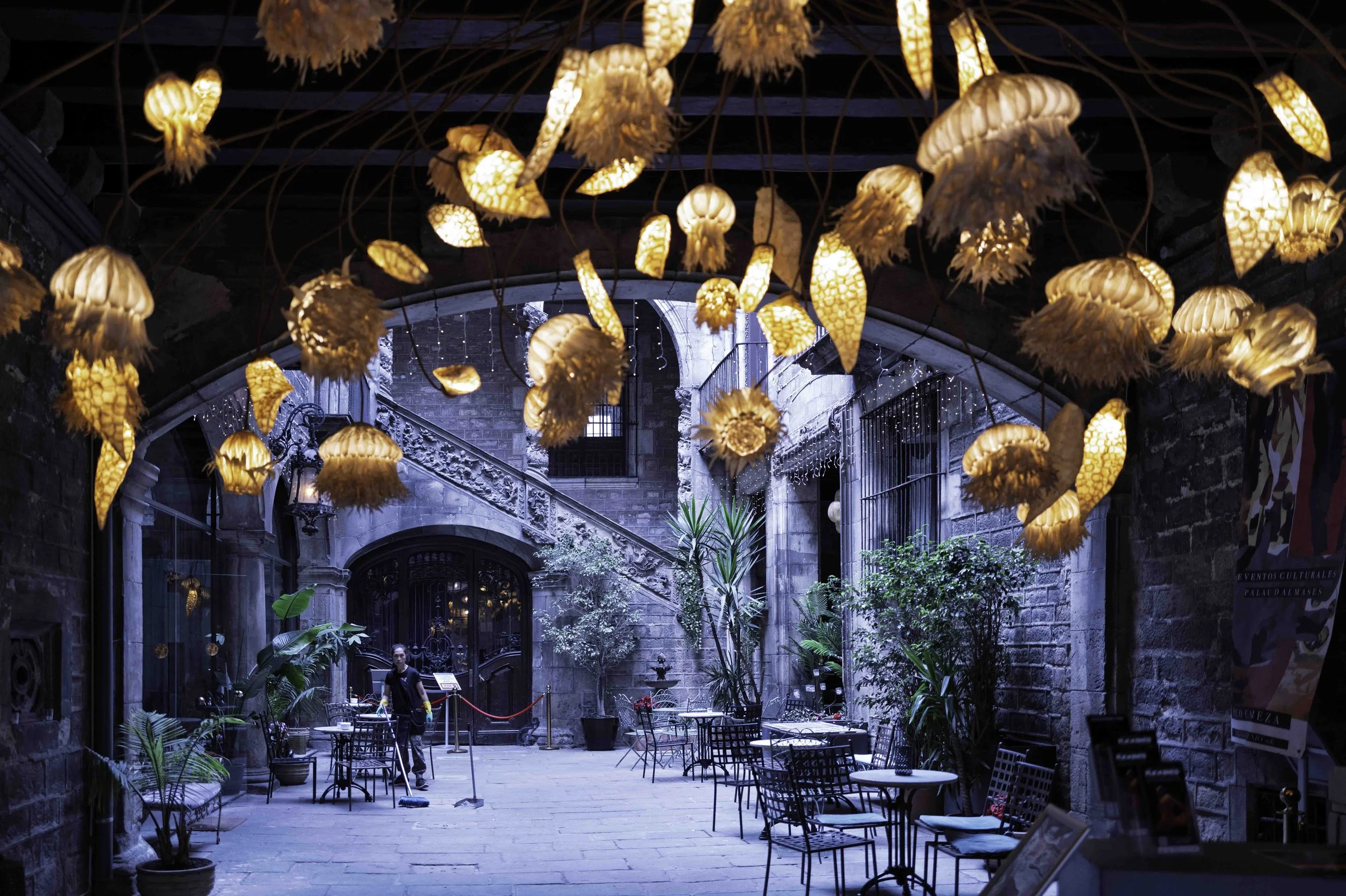 Glowing jellyfish lanterns hanging in the courtyard of Palau del Lloctinent, Gothic Quarter Barcelona, with a lone worker sweeping the flagstones below — Leica Q3