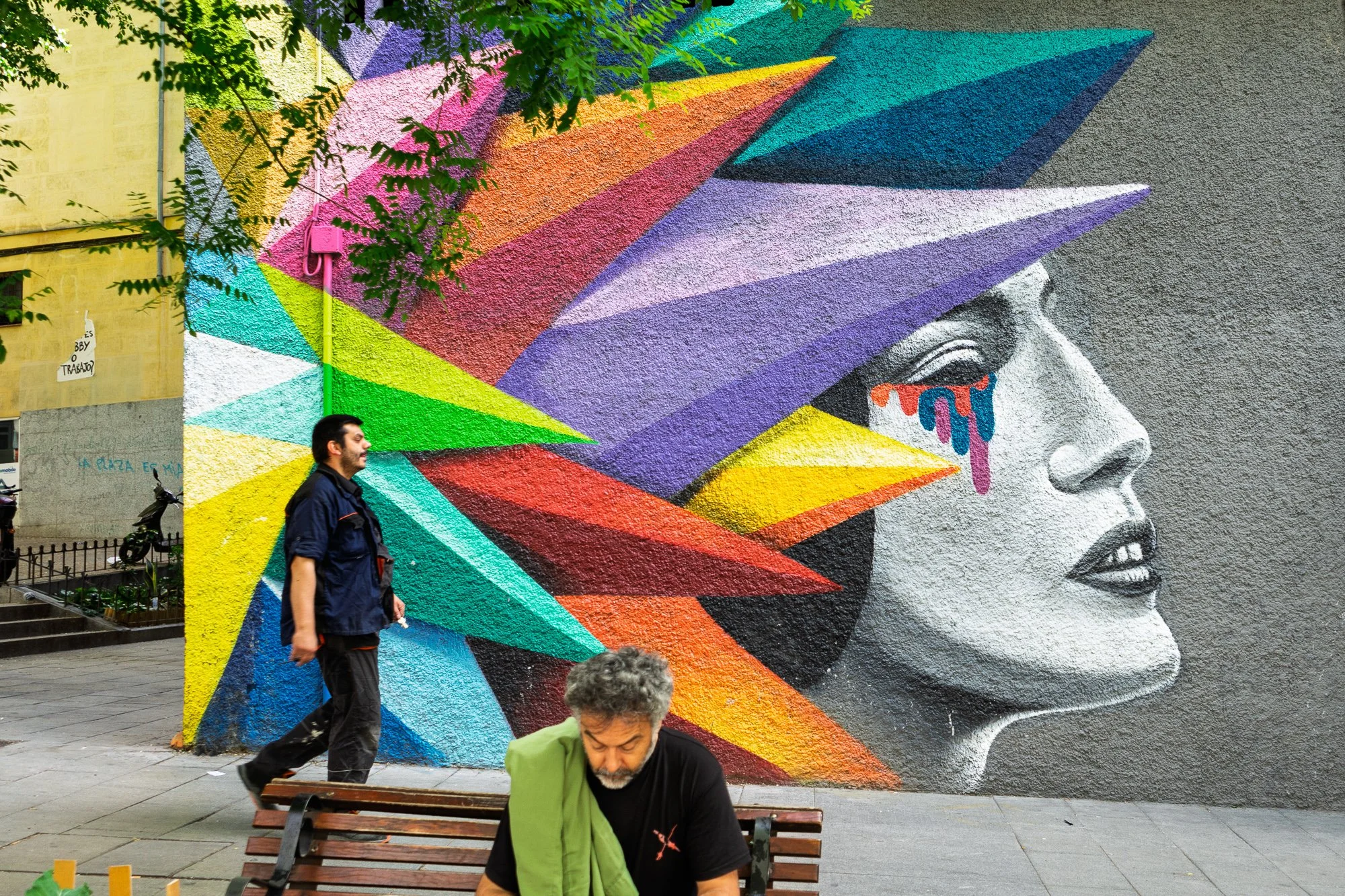 A man walks past the same Lavapiés mural while another sits on a bench in front of it, a towel over his shoulder