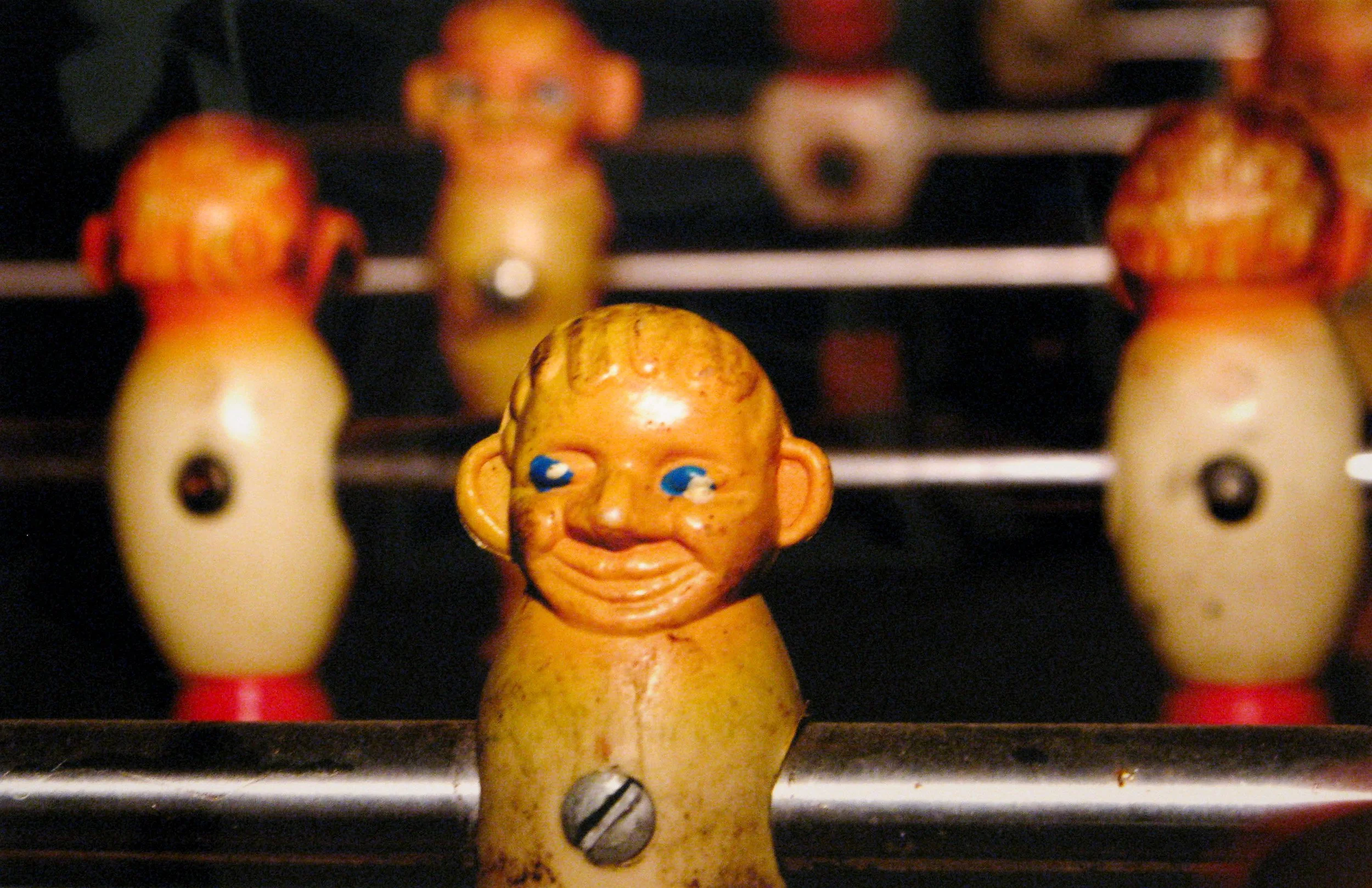 Futbolí, a photograph by Cosme Lapena: close-up of a worn wooden table football player with painted blue eyes and a wide grin, other players blurred behind.
