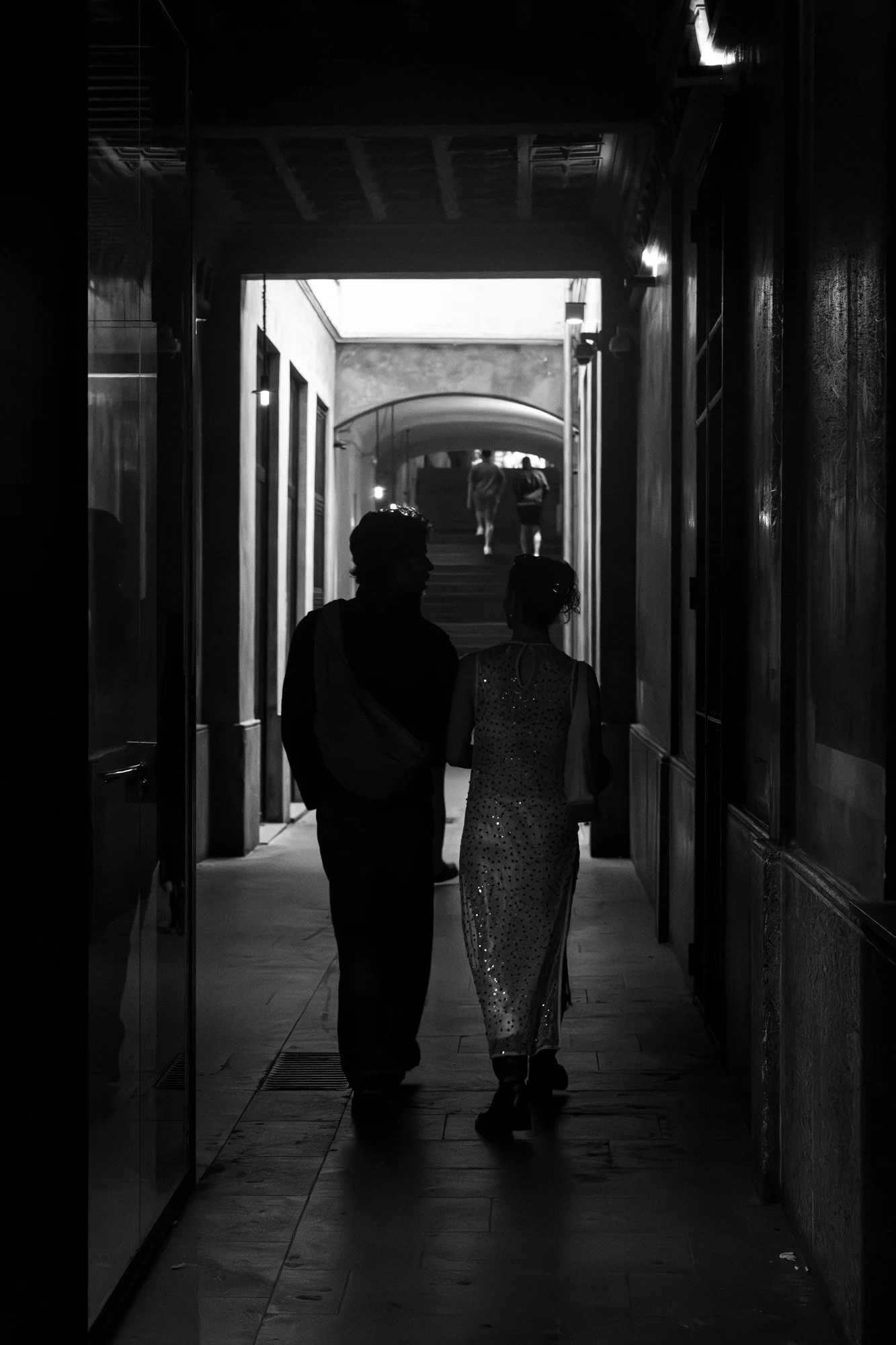 Couple silhouetted in a dark Gothic Quarter passageway at night, Barcelona, lit arch visible in the background — Sony a7R IV