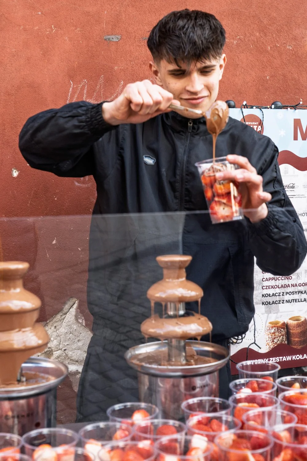 Young vendor pouring melted chocolate over a cup of strawberries beside chocolate fountains, Warsaw — Sony a7R IV