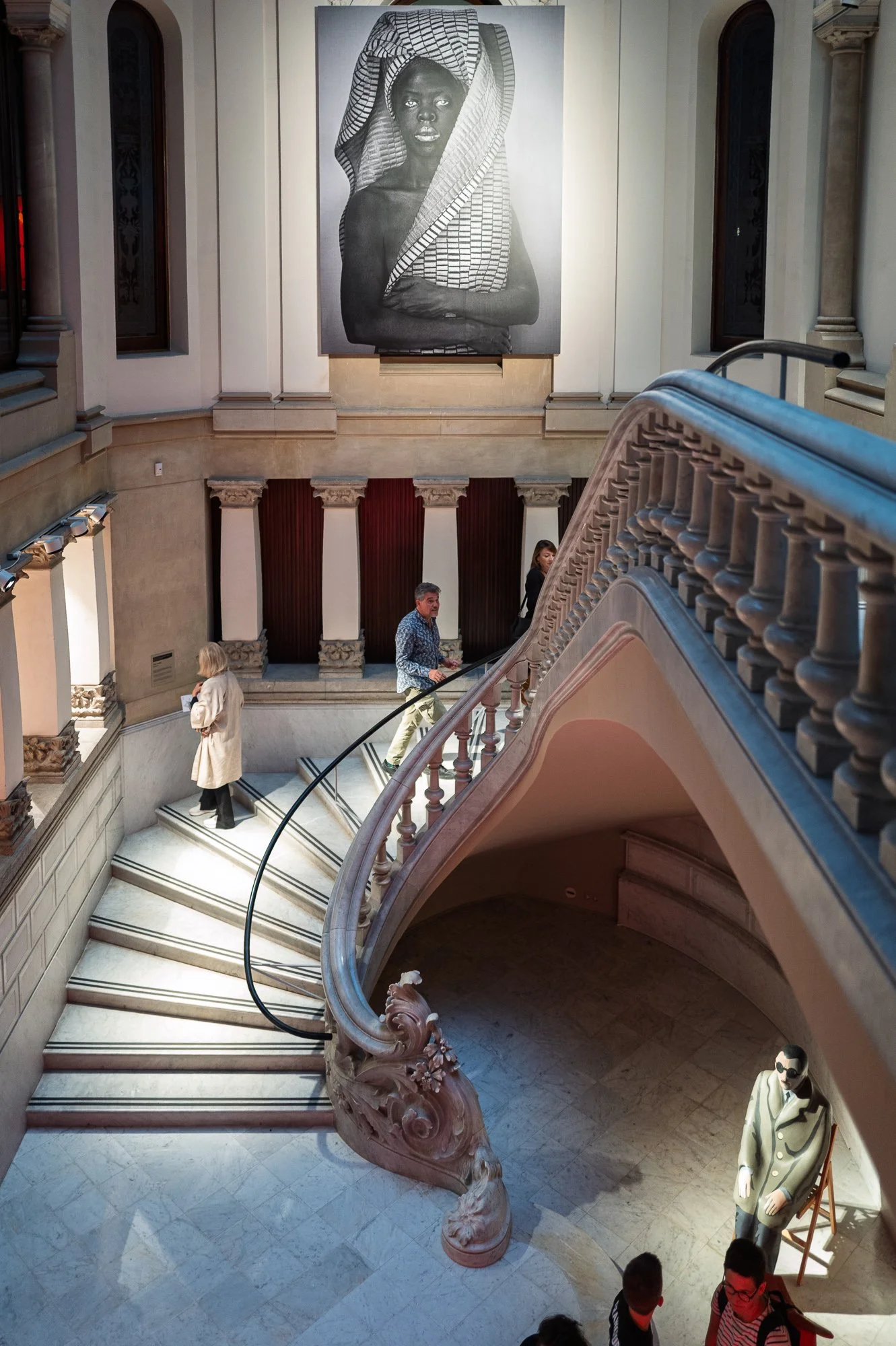 Visitors on a curving marble staircase below a huge black and white portrait of a woman, the seated figure at bottom right