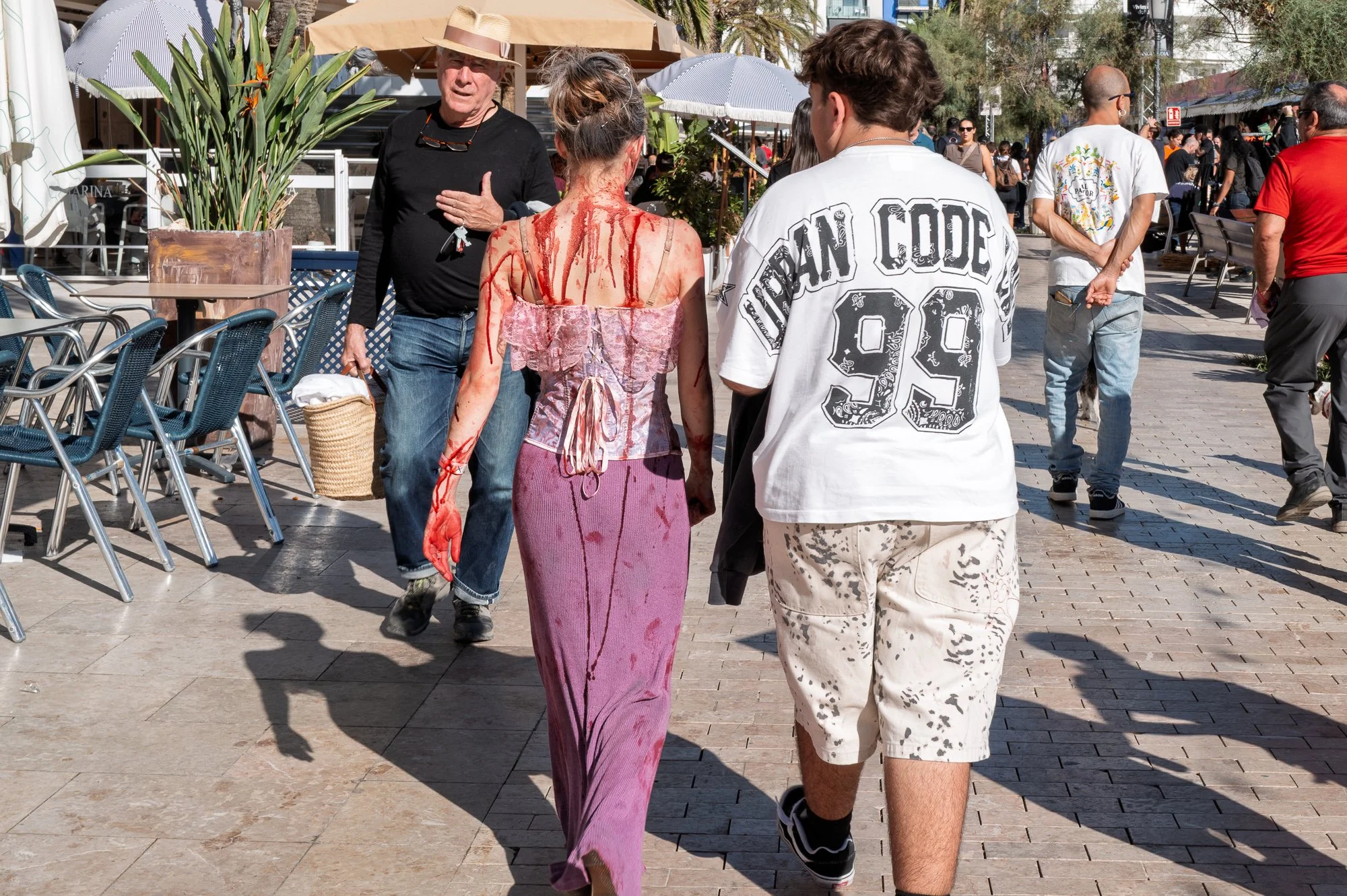 Sitges Zombie Walk: woman with blood-streaked back walks the seafront past an older man in a straw hat — Leica Q3