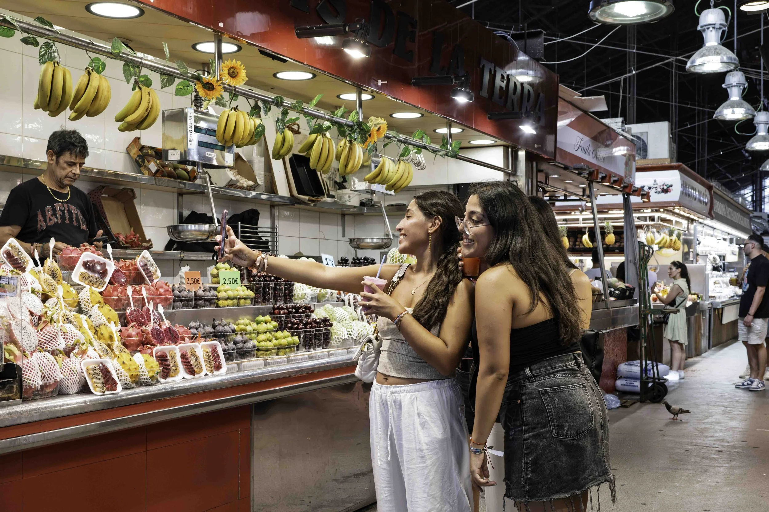 Two young women taking a selfie at a colourful fruit stand inside La Boqueria market Barcelona while the vendor looks on — Leica Q3