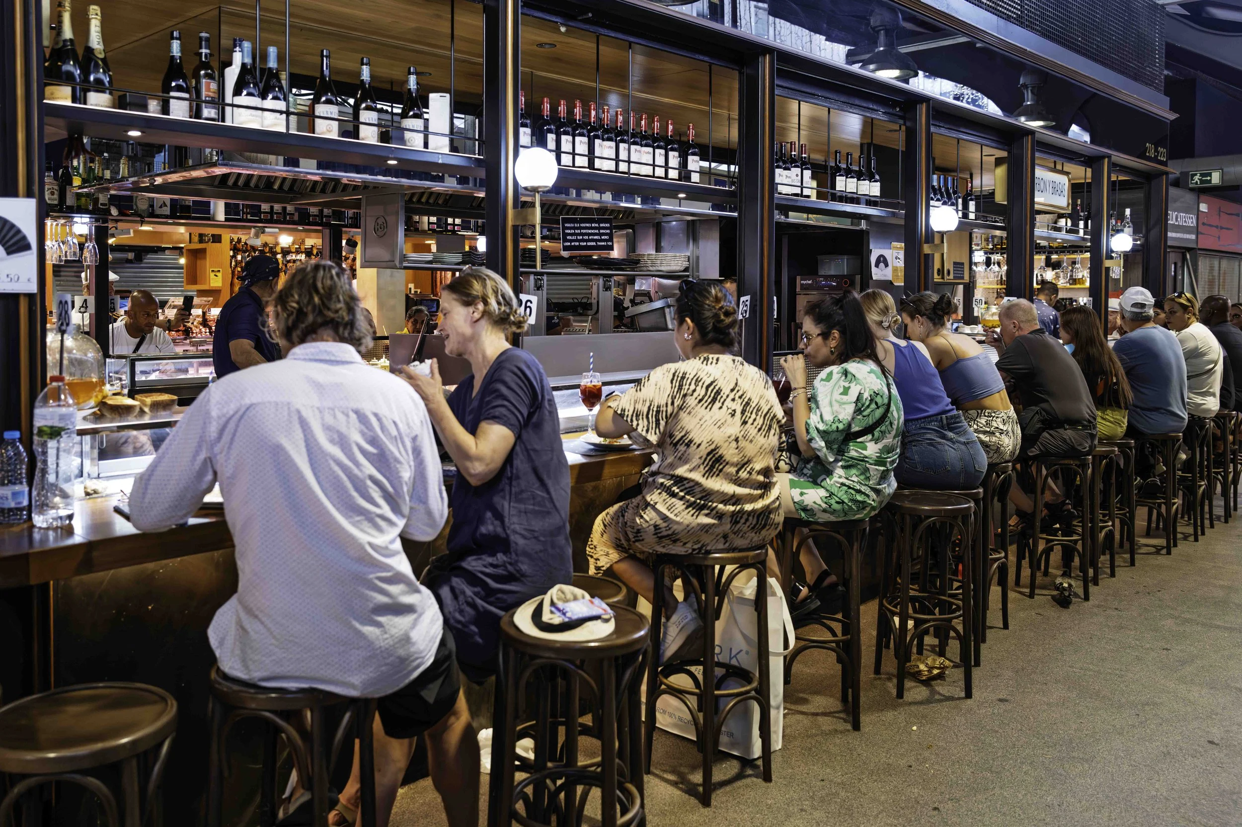 Customers sitting shoulder to shoulder on bentwood stools along a busy bar counter inside La Boqueria market Barcelona — Leica Q3