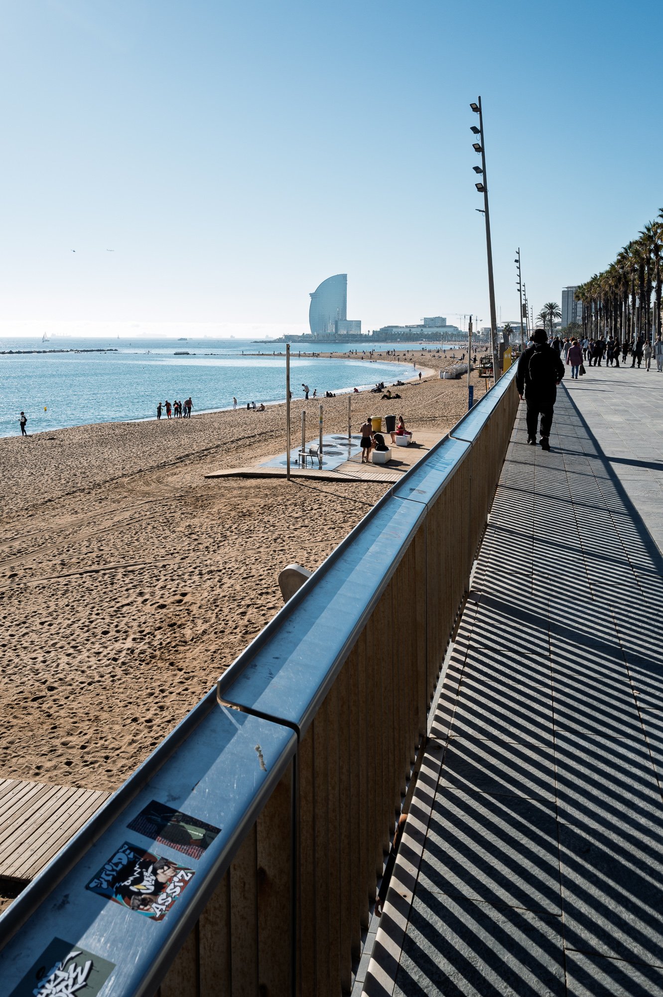 Promenade railing casting striped shadows toward the W hotel, Barceloneta beach — Leica Q3