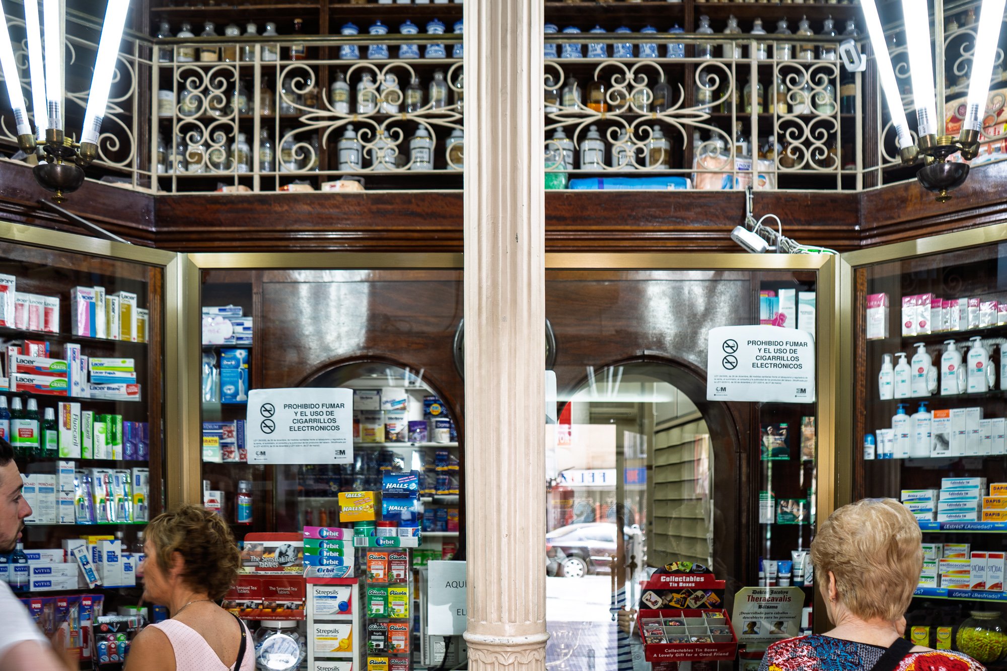 Old pharmacy interior with porcelain jars on wooden shelves above modern products and customers at the counter