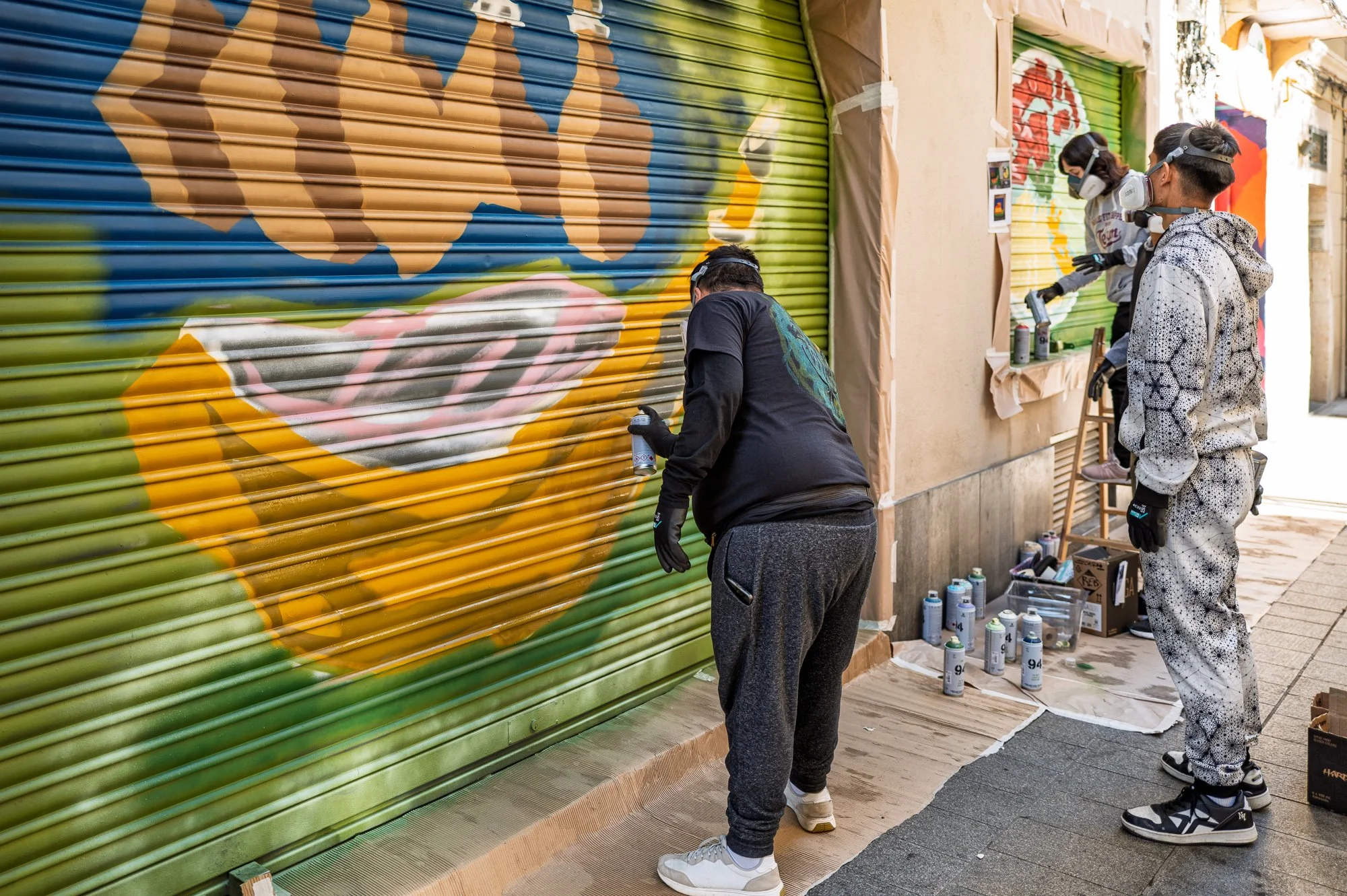 Street artists in respirator masks spray-painting shop shutters in a narrow Barceloneta street — Leica Q3