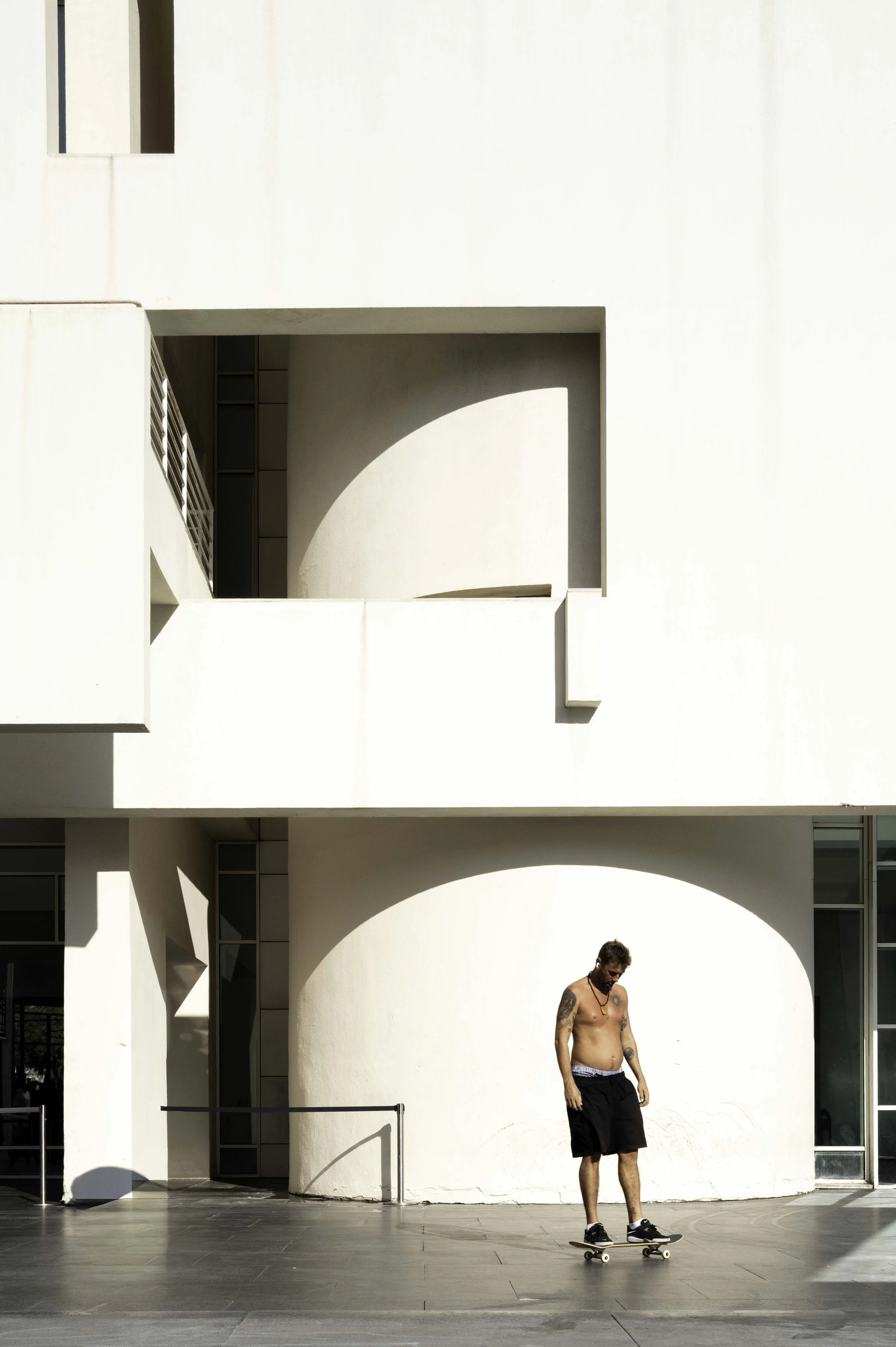 Shirtless skateboarder standing on a skateboard beneath a white arched facade at MACBA Barcelona in summer — Leica Q3