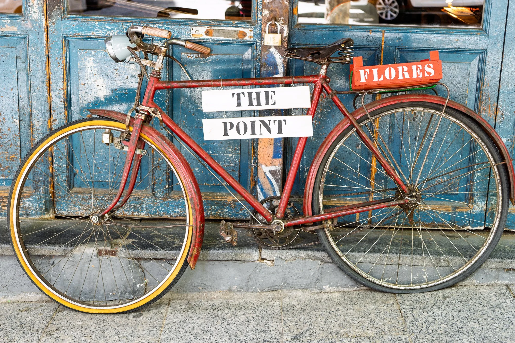 Old red bicycle with a crate marked Flores and a sign reading The Point, leaning on peeling blue doors, Madrid