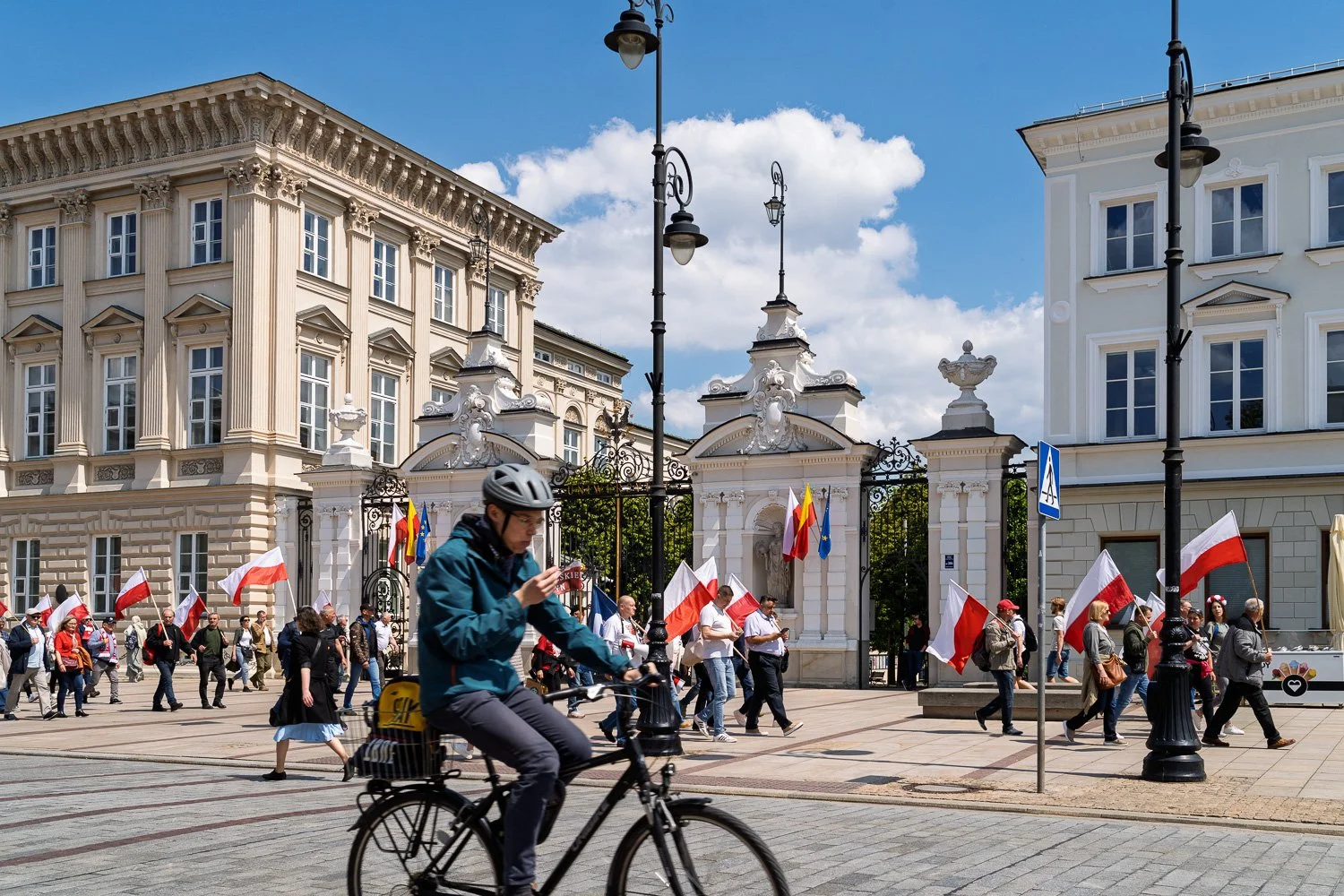 Cyclist on his phone in front of the University of Warsaw gate as marchers pass with flags — Sony a7R IV