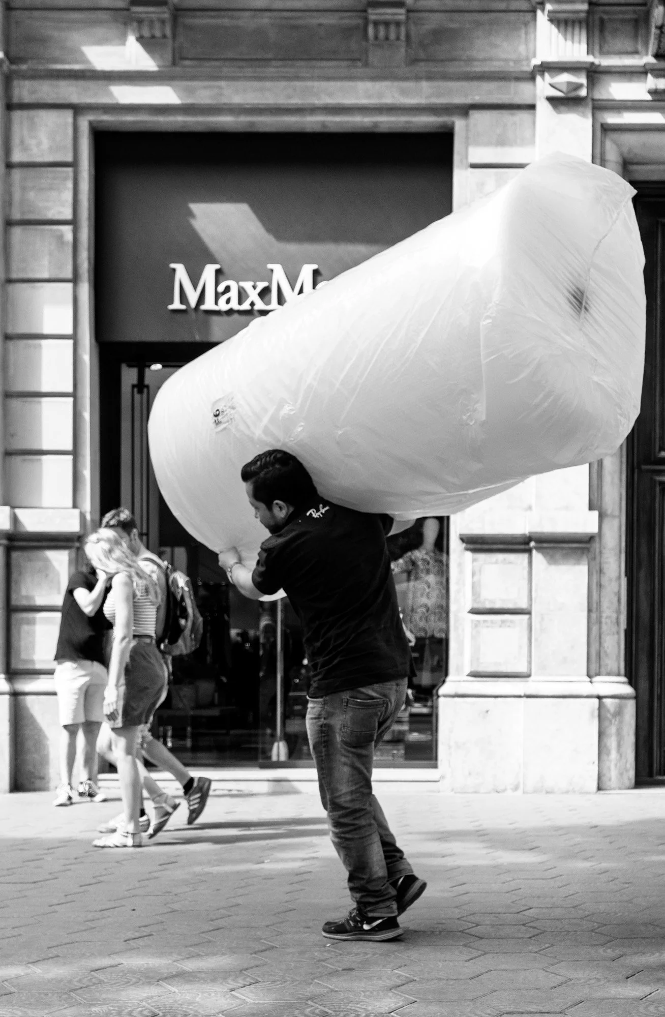 Man carrying an oversized plastic-wrapped package on his shoulder past a luxury shop in Barcelona street photography — Sony a7R IV