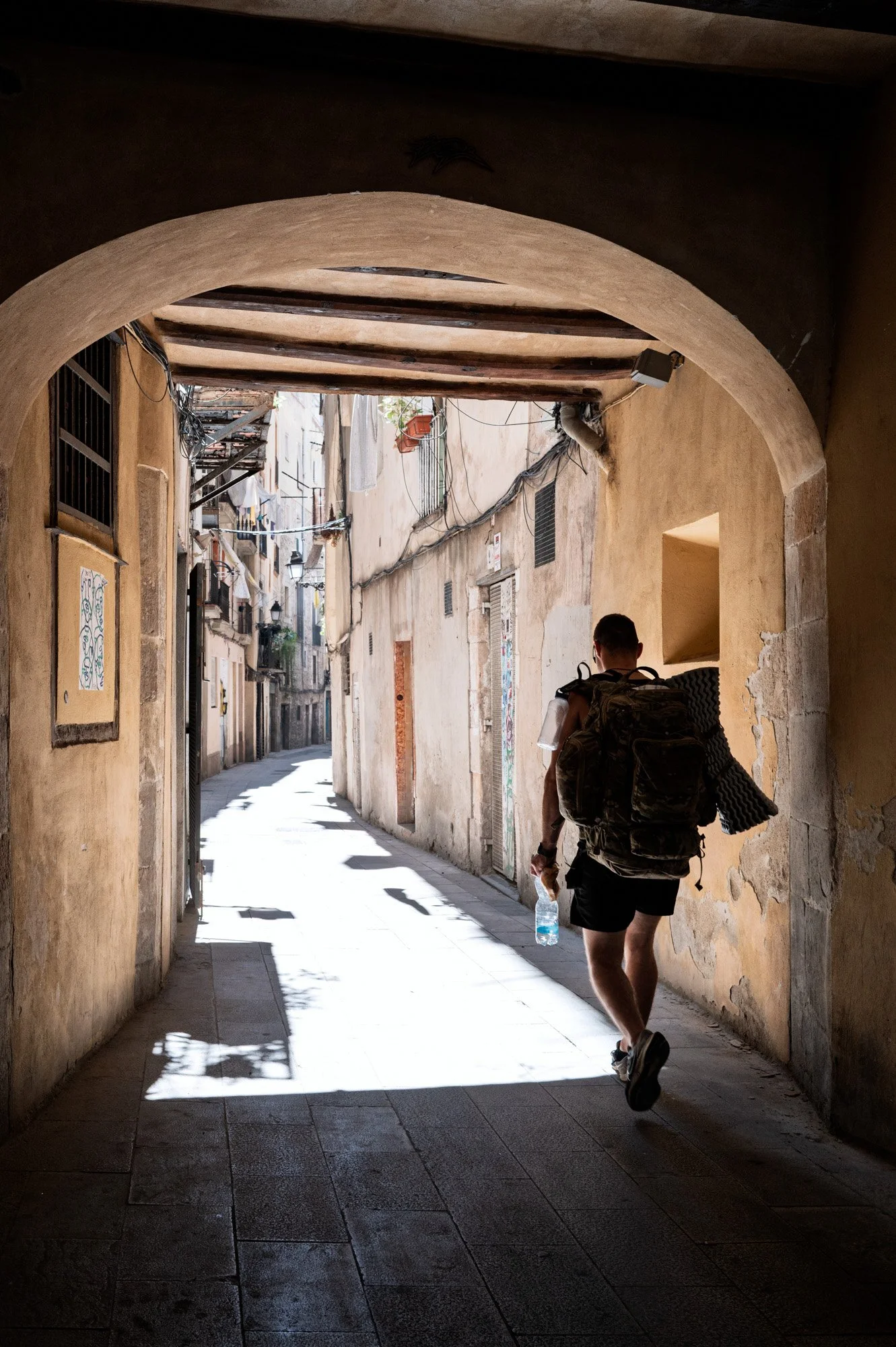 Backpacker walking from shadow into sunlight under a stone archway, El Born — Leica Q3