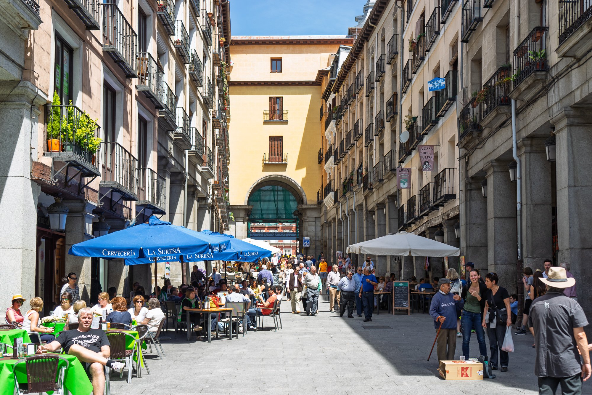 Busy street of terraces under blue umbrellas leading to the yellow Arco de Toledo of the Plaza Mayor, Madrid