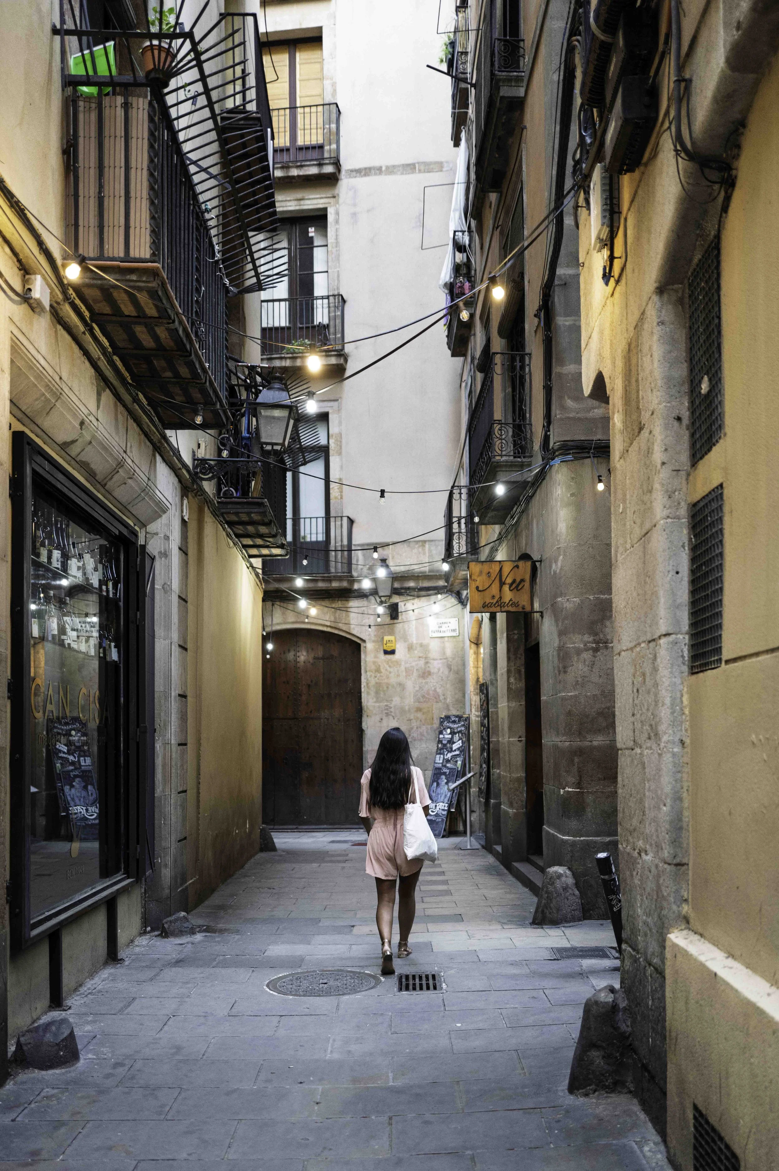 Woman walking alone down a narrow Gothic Quarter Barcelona alley at night lit by warm string lights overhead — Leica Q3
