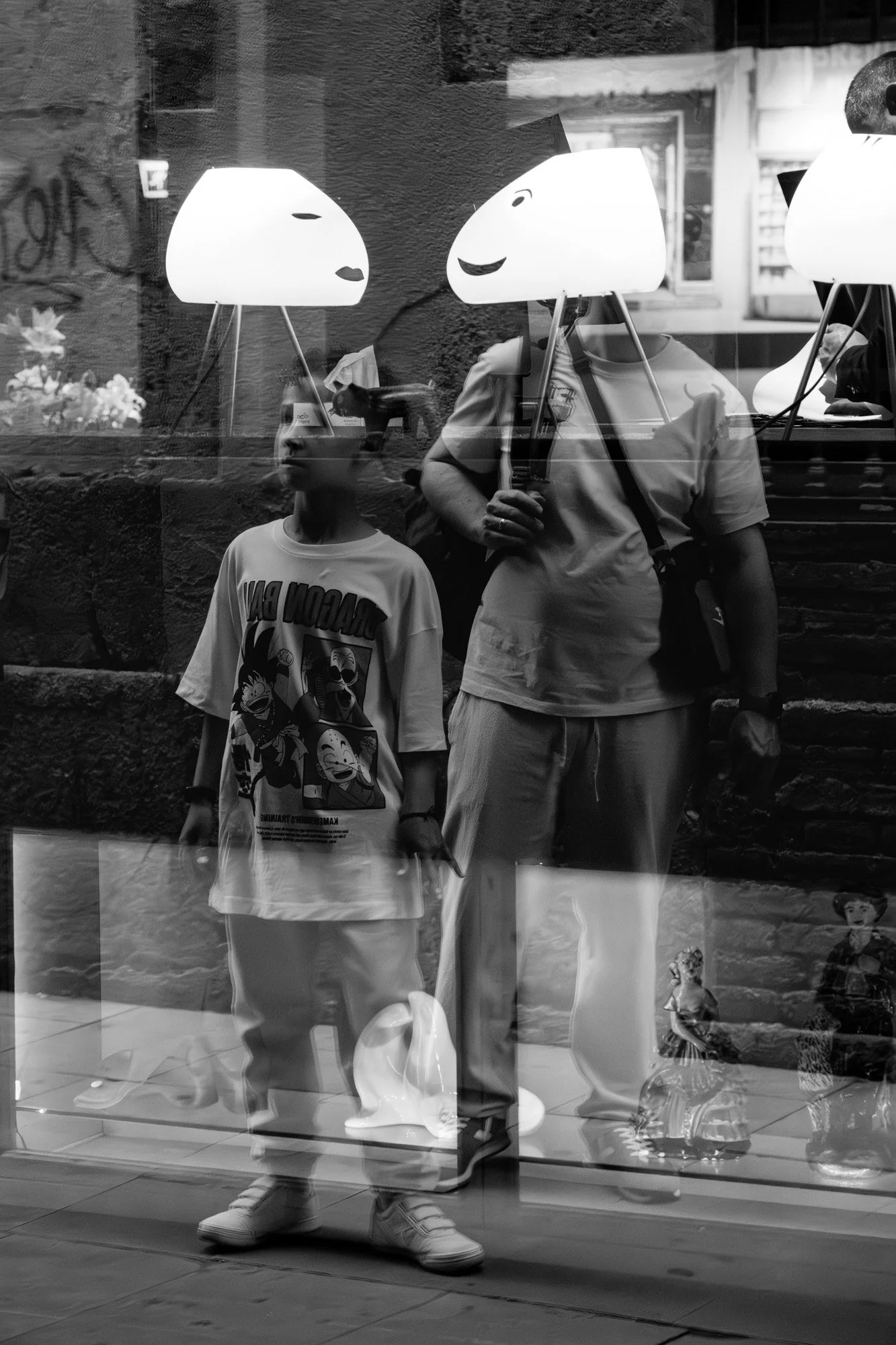 Father and son outside MACBA Barcelona design shop, with displayed lamp shades appearing in window reflection as heads on their shoulders — Sony a7R IV