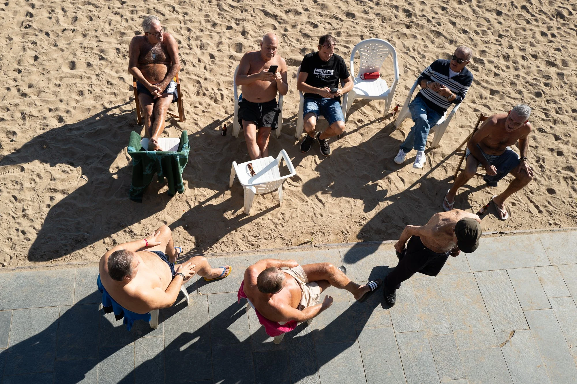 Neighbors talking in plastic chairs on the sand, seen from the promenade, Barceloneta — Leica Q3