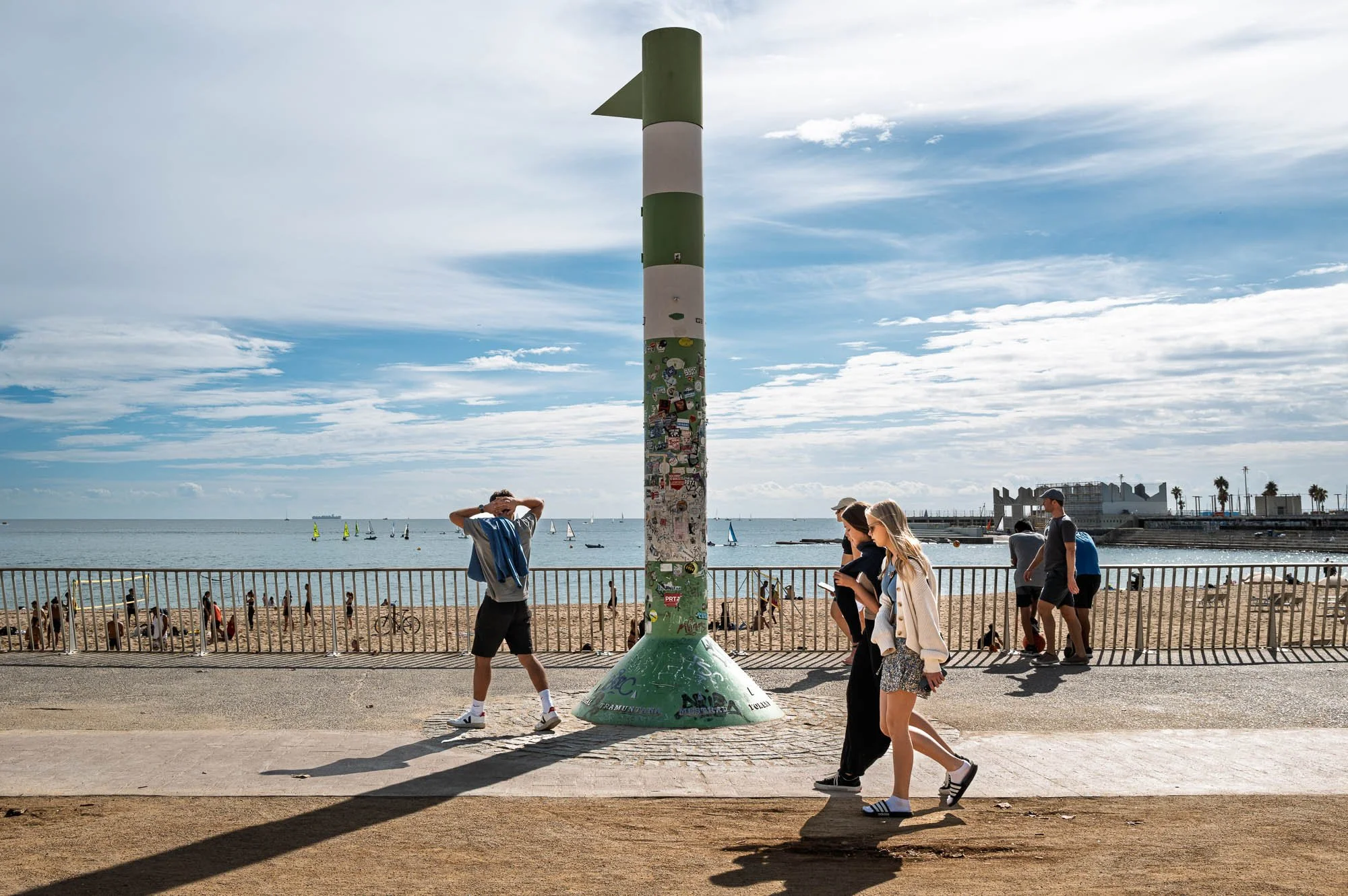 People walking past a green and white beacon on the Barceloneta seafront, beach and sailboats behind — Leica Q3
