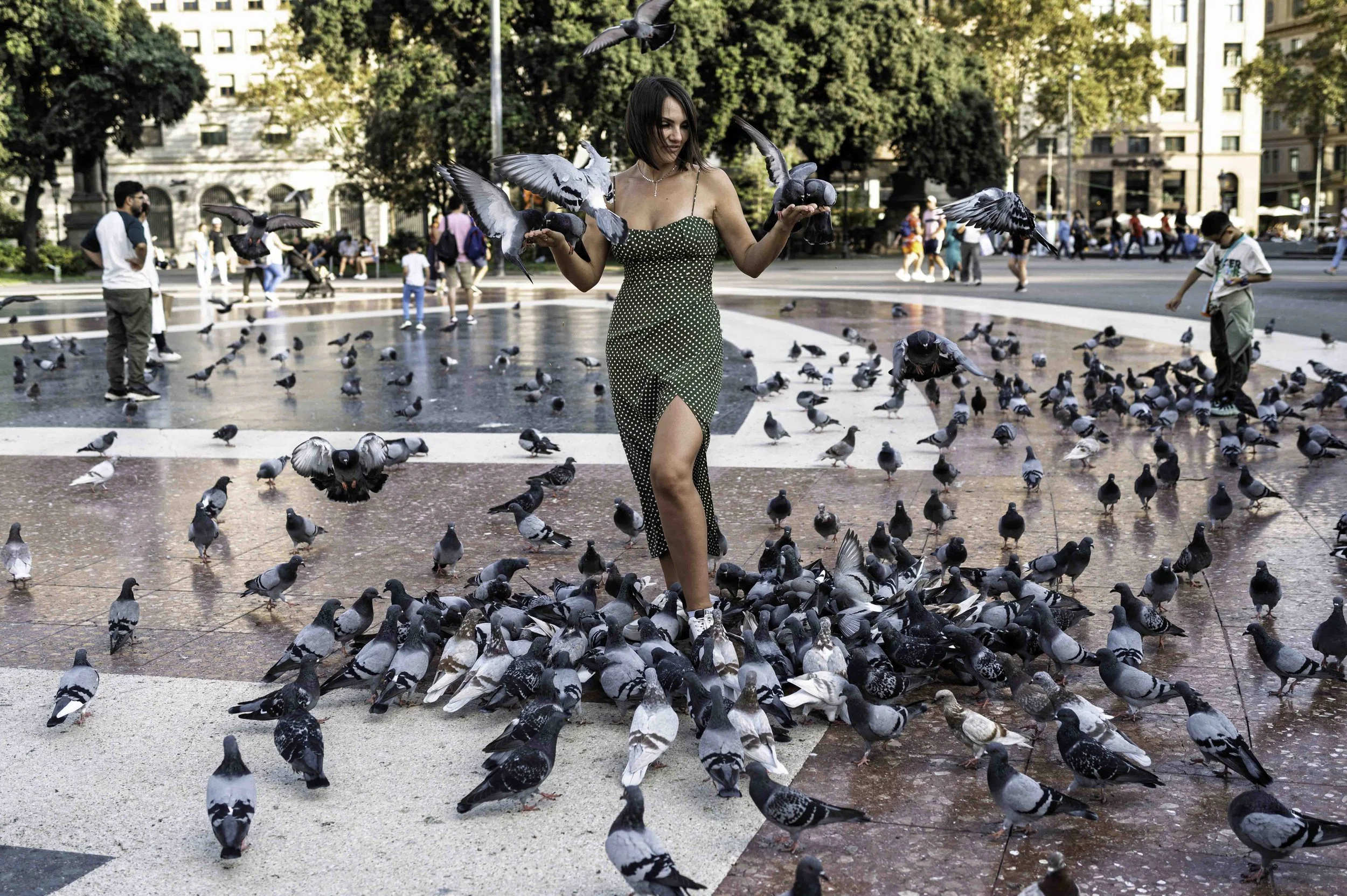 Young woman in a green polka-dot dress feeding pigeons with outstretched arms at Plaça Catalunya Barcelona surrounded by a large flock — Leica Q3