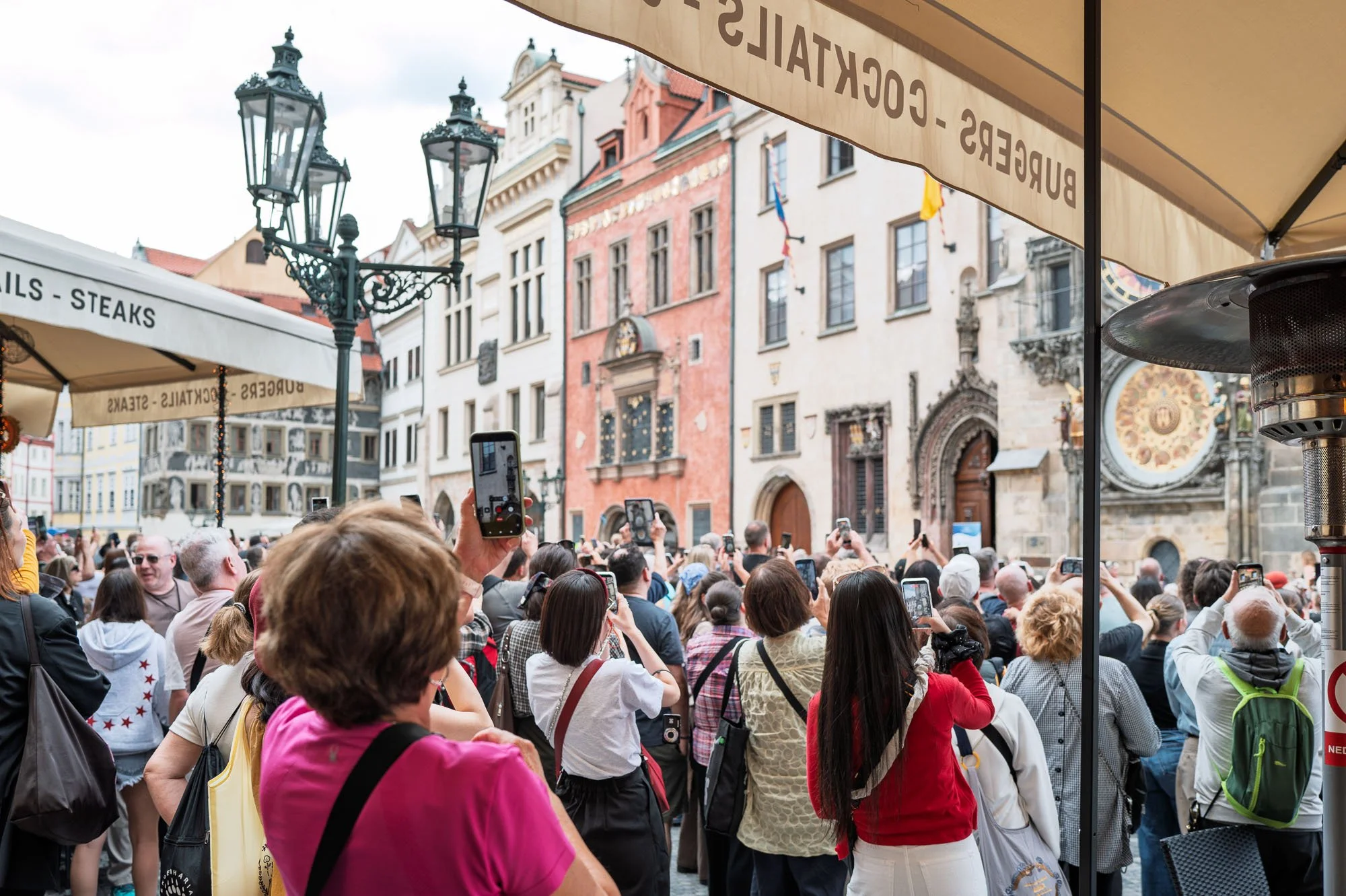 Raised phones filming the Astronomical Clock from under a terrace umbrella, Old Town Square, Prague — Leica Q3