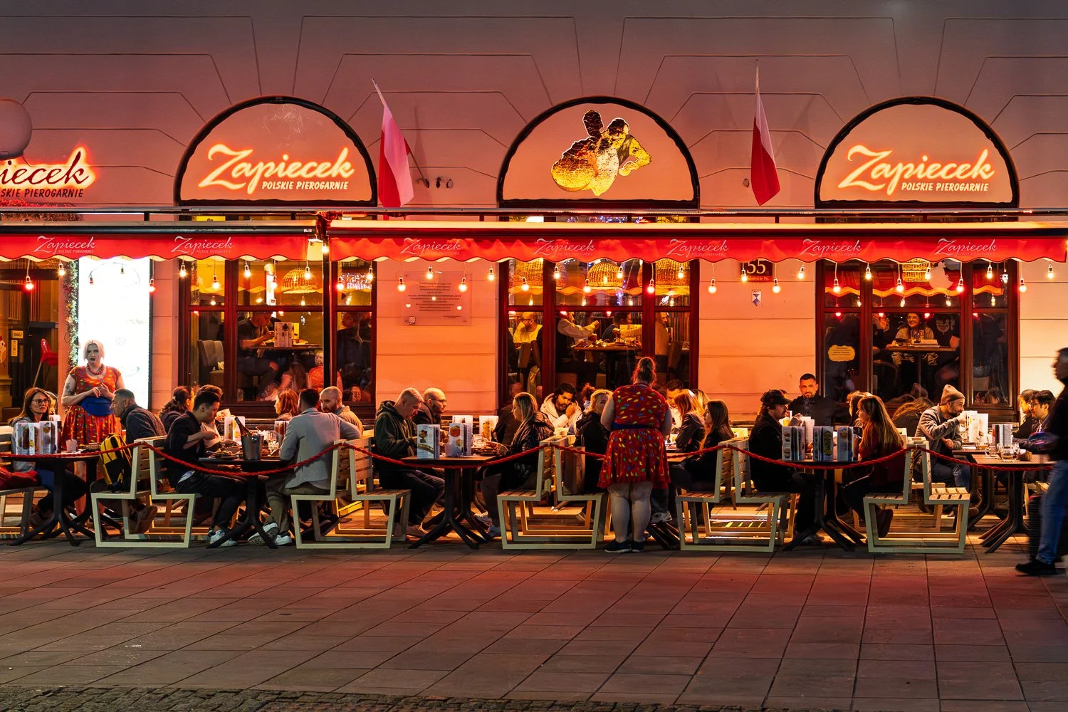 Zapiecek pierogi restaurant glowing red at night, waitresses in dotted dresses on a full terrace, Warsaw — Sony a7R IV