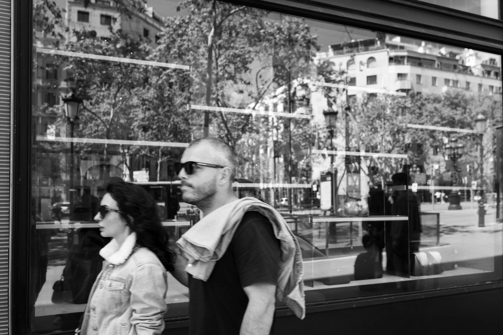 Couple walking past a shop window reflection on Passeig de Gràcia Barcelona, reflections layered with boulevard trees — Leica Q3