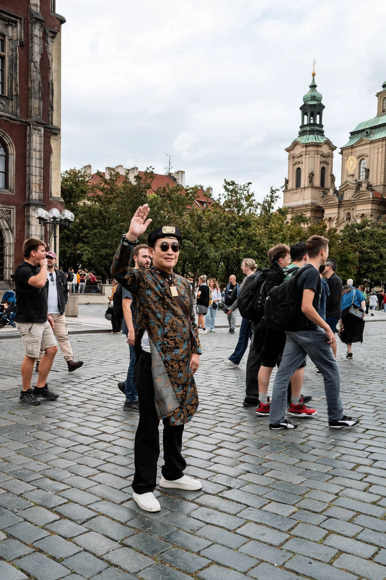 Man in a patterned coat waving at the camera, St Nicholas Church behind, Old Town Square, Prague — Leica Q3