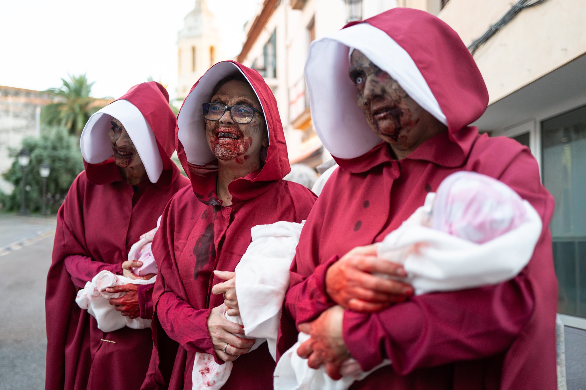 Three zombie handmaids in red cloaks and white bonnets carrying bloodied baby dolls, Sitges — Leica Q3