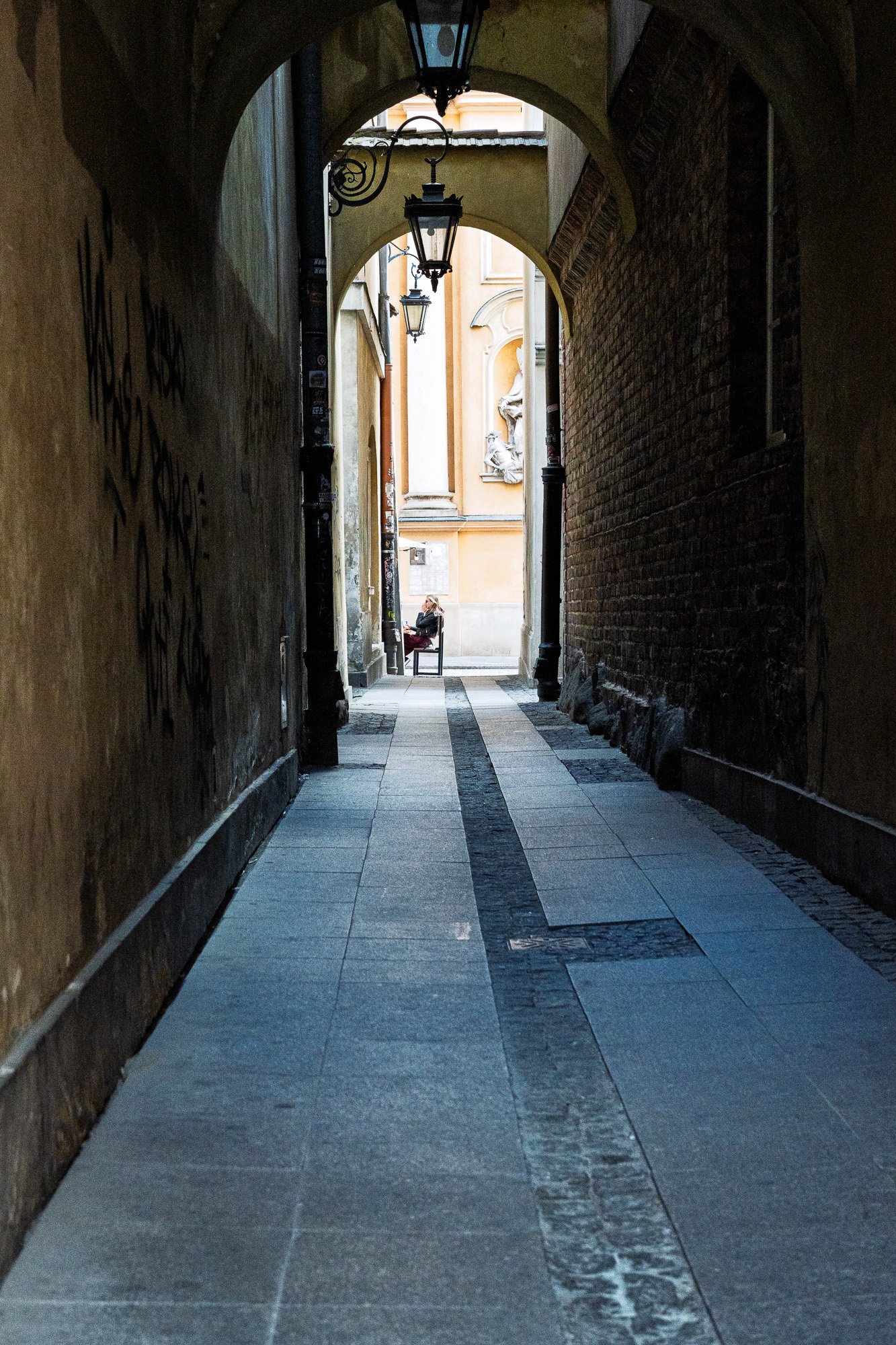 The same Old Town passage in Warsaw, now with a woman on the bench in sunlight at the far end — Sony a7R IV