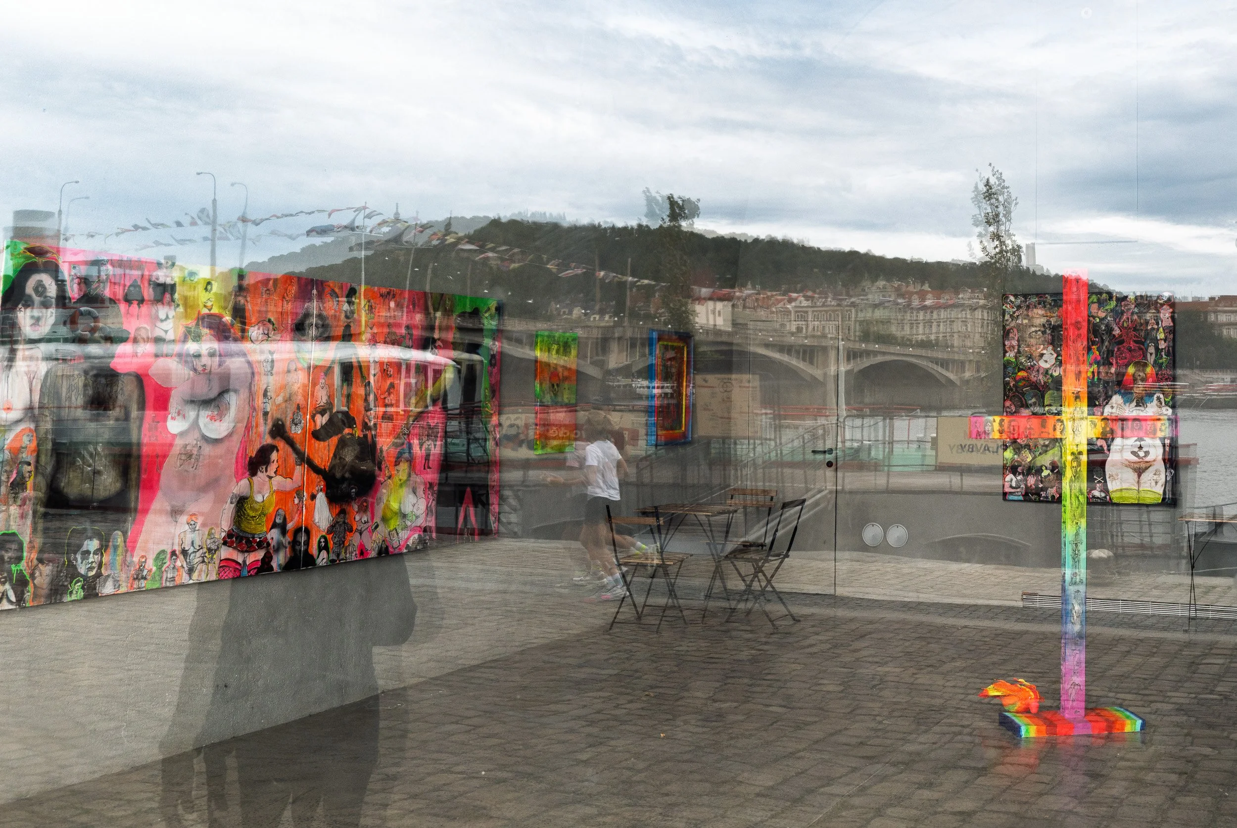 Gallery window with bright paintings, Vltava bridge and a runner reflected, Prague reflections — Leica Q3