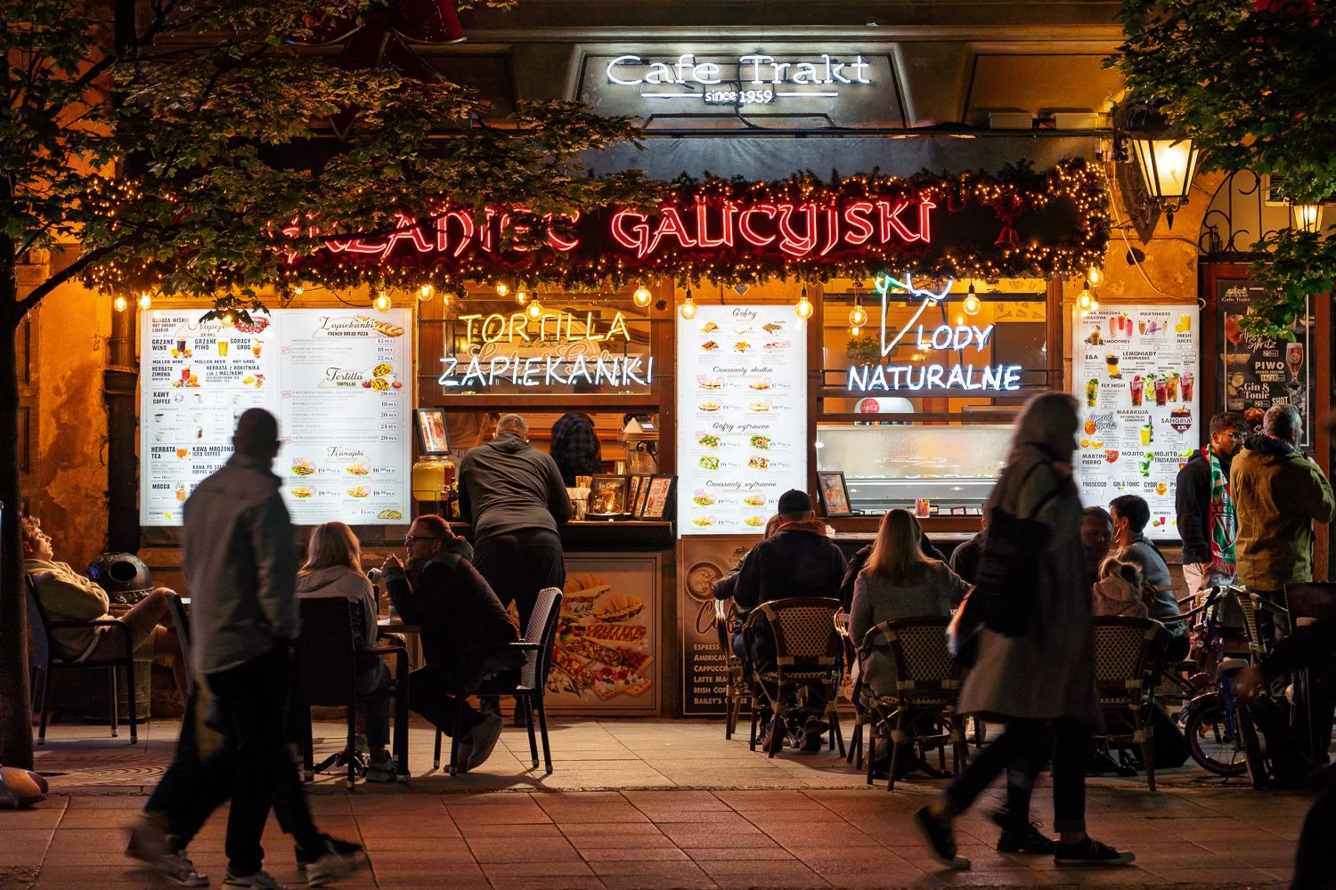 Cafe Trakt at night with neon signs, terrace tables and blurred passers-by on the Warsaw Royal Route — Sony a7R IV